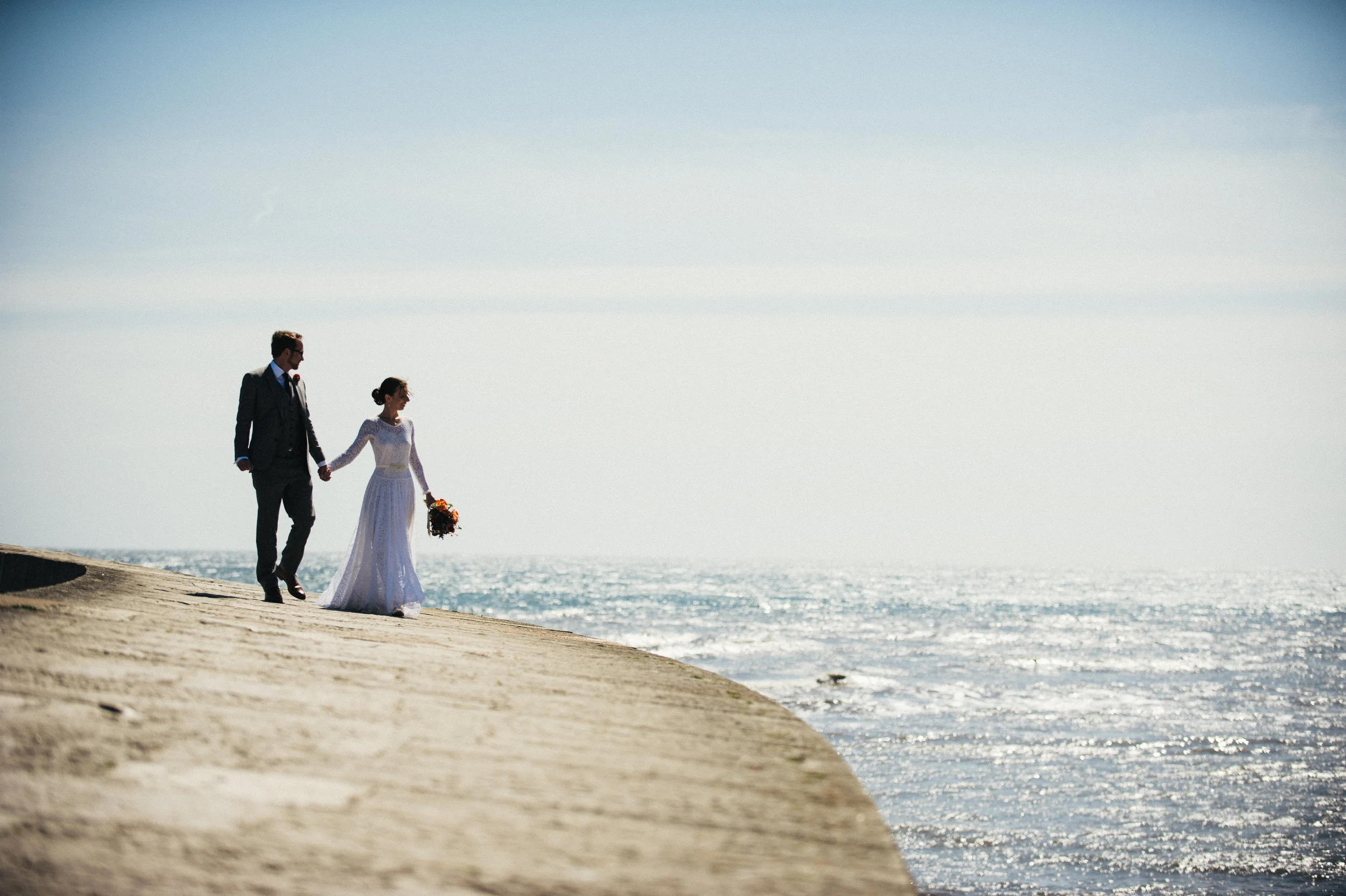 A bride and groom walk hand in hand along a beach shoreline with the ocean in the background, on a sunny day.