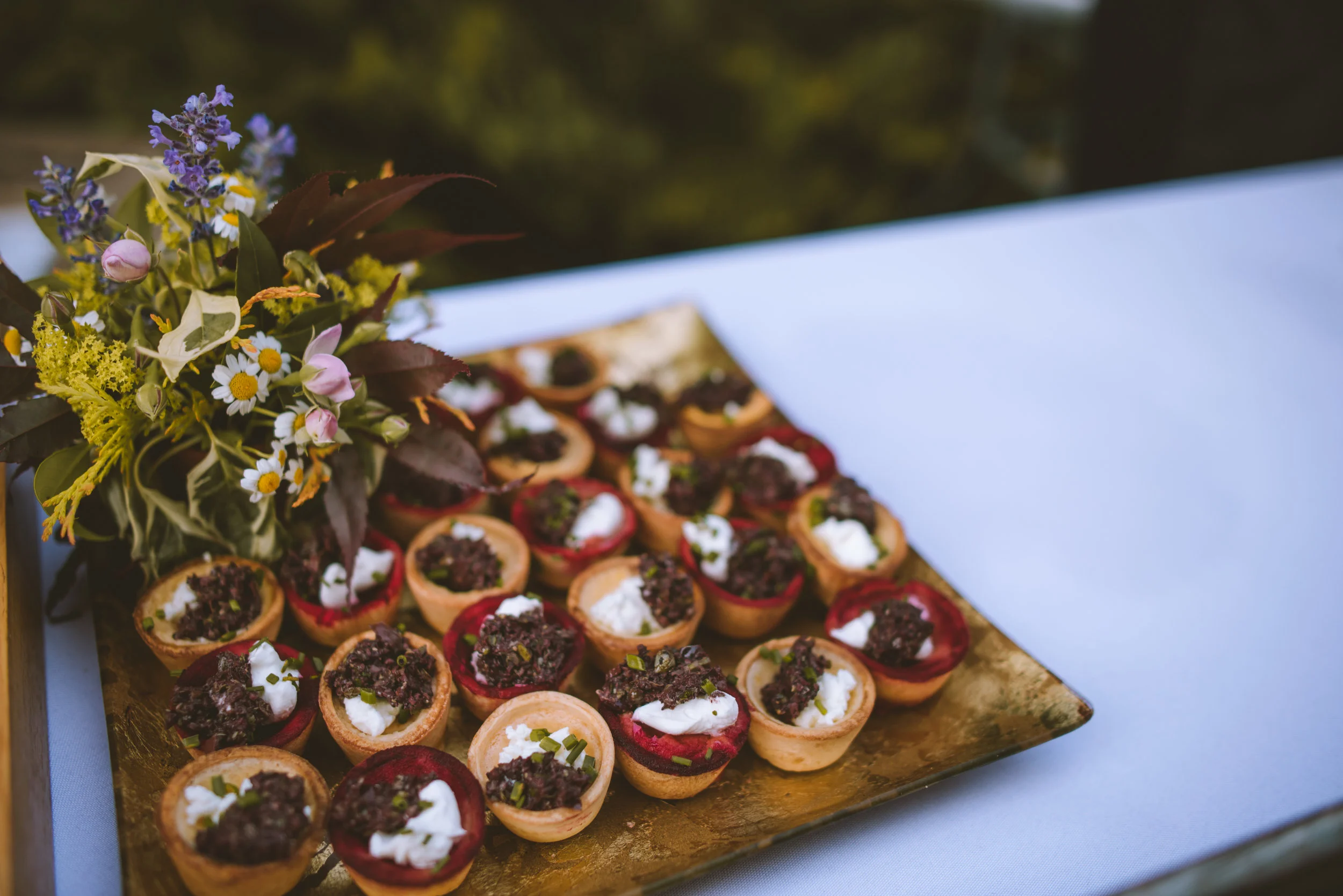 An assortment of bite-sized appetizers arranged on a golden tray, garnished with herbs and topped with cream and dark toppings, next to a small bouquet of mixed flowers.