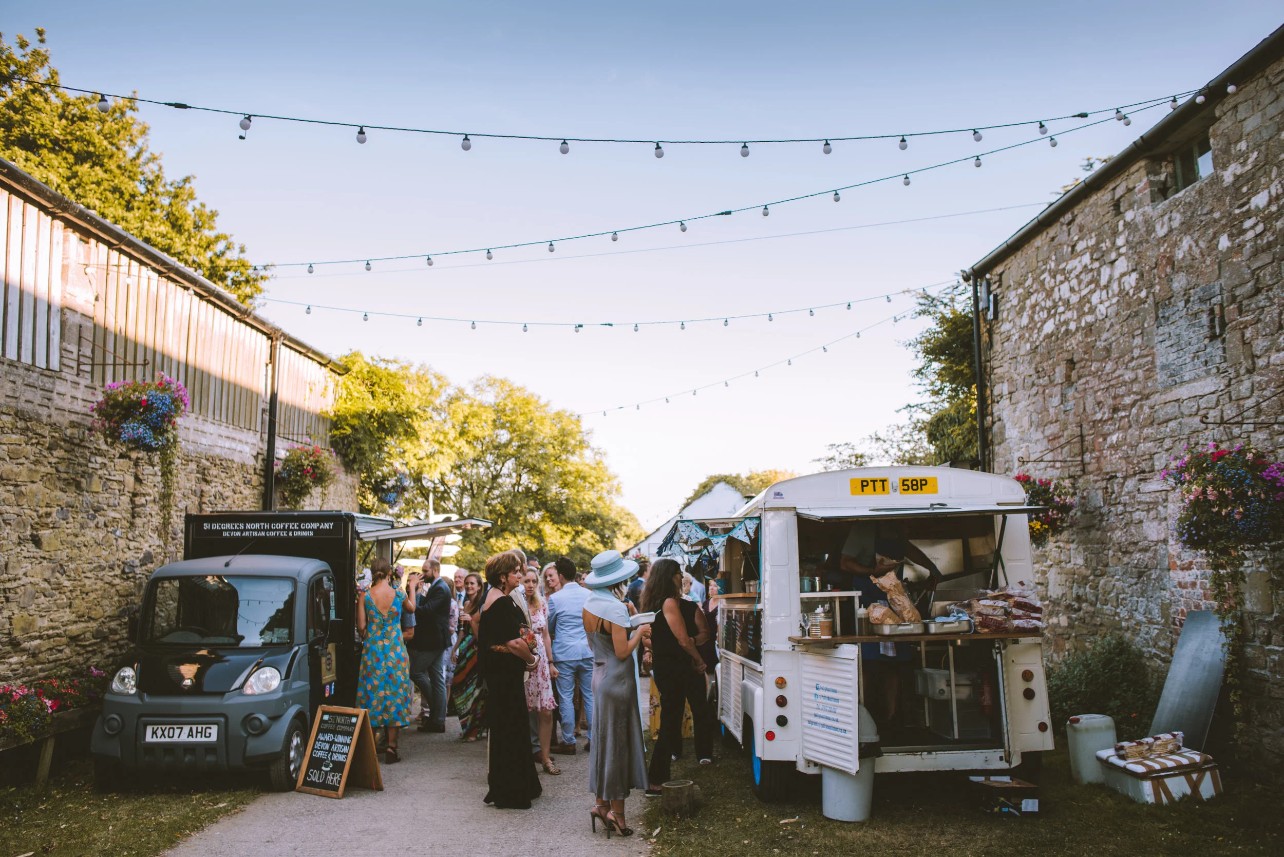 People gather at an outdoor food truck event on a sunny day, with string lights overhead and stone buildings lining the sides.