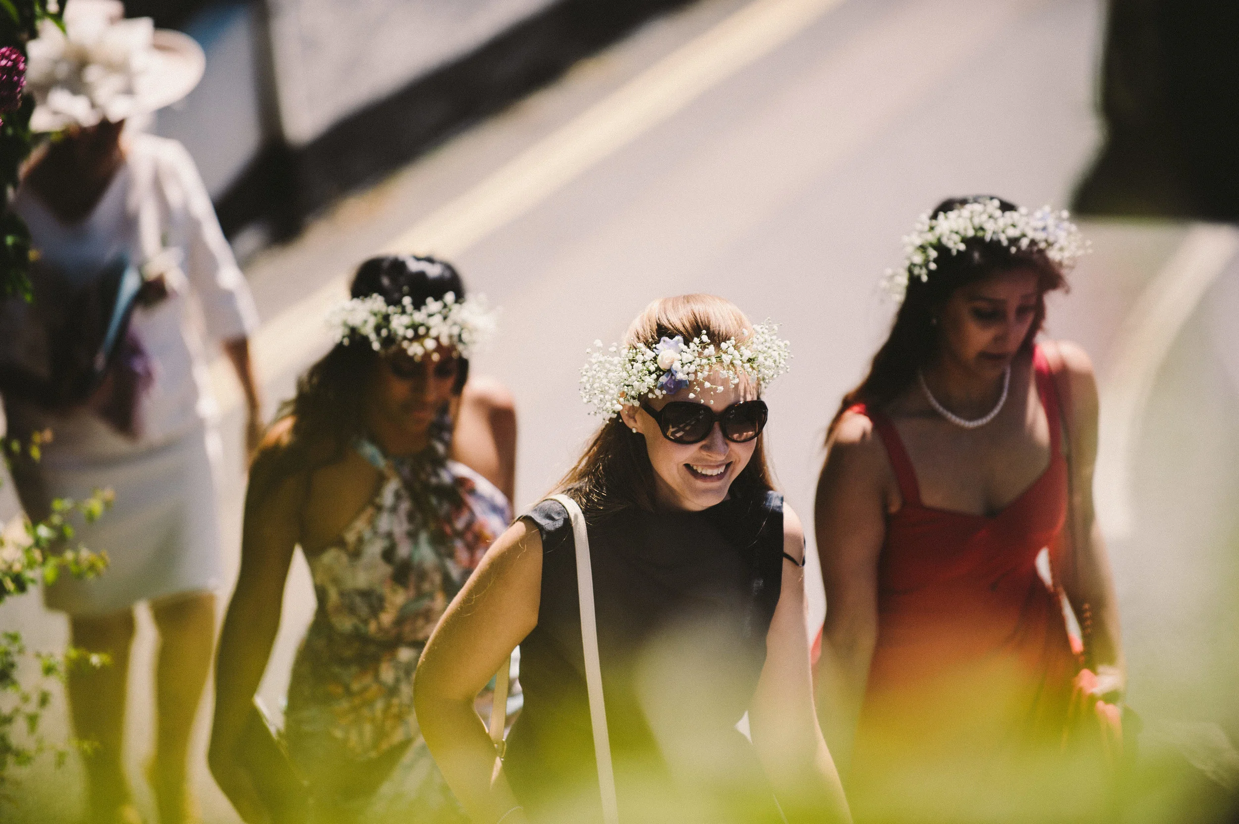 A group of women dressed in summer attire walking outdoors, wearing flower crowns, and smiling. One woman is wearing sunglasses and a black dress, while others are in colorful dresses.