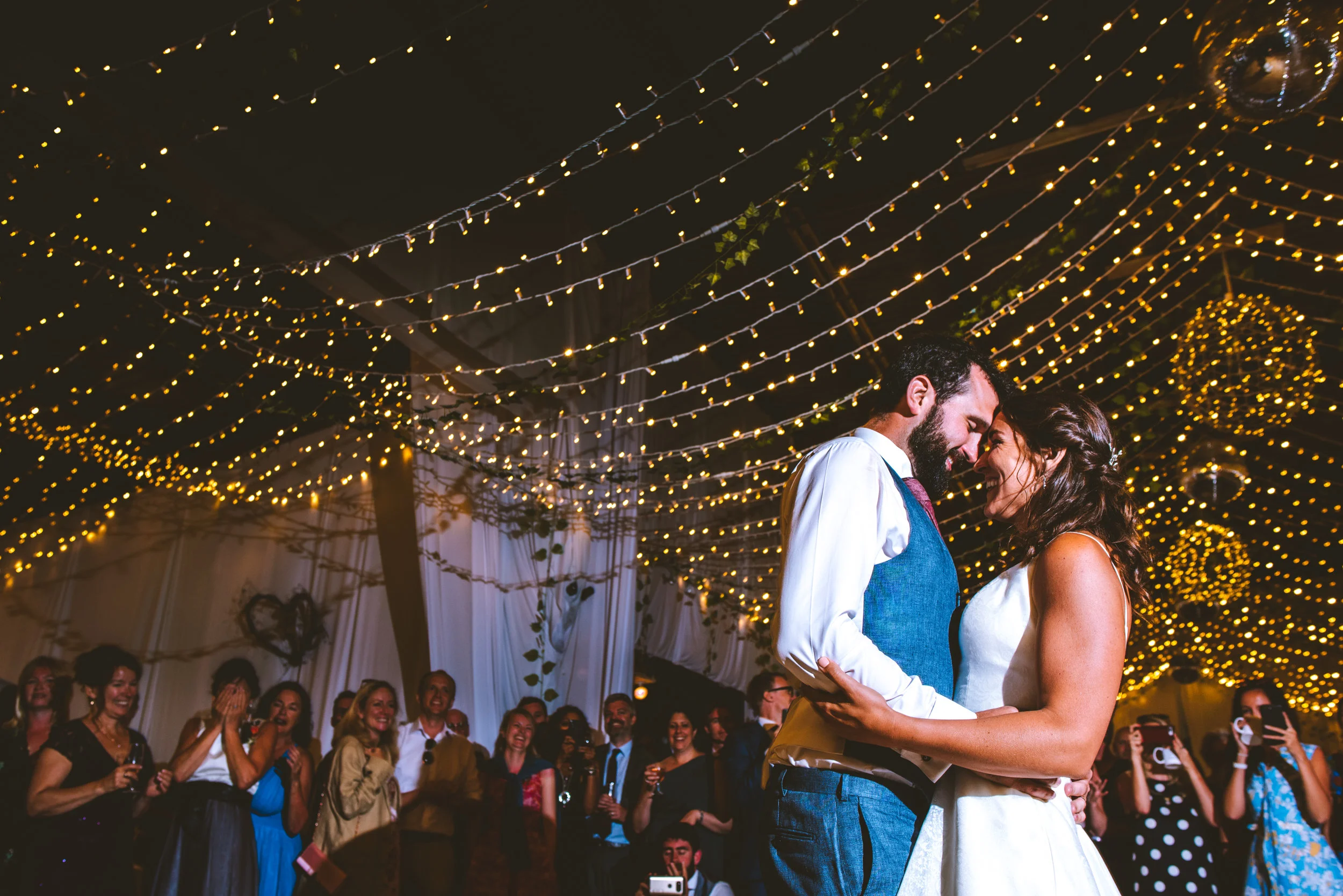A bride and groom dancing at their wedding reception, surrounded by guests, under hanging string lights and chandeliers.