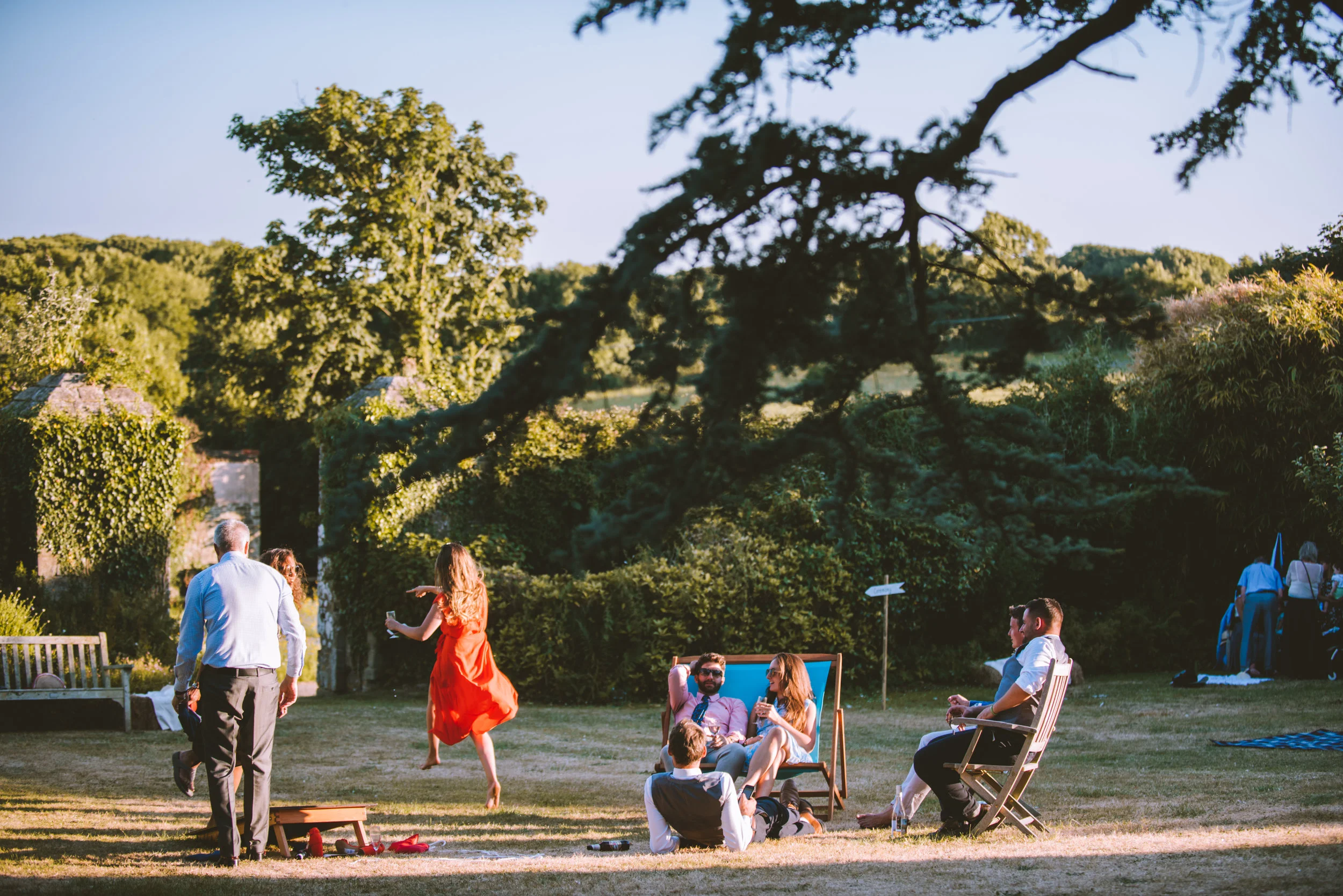 People enjoying a garden party outdoors in a lush green area with trees and bushes, some sitting and others dancing.