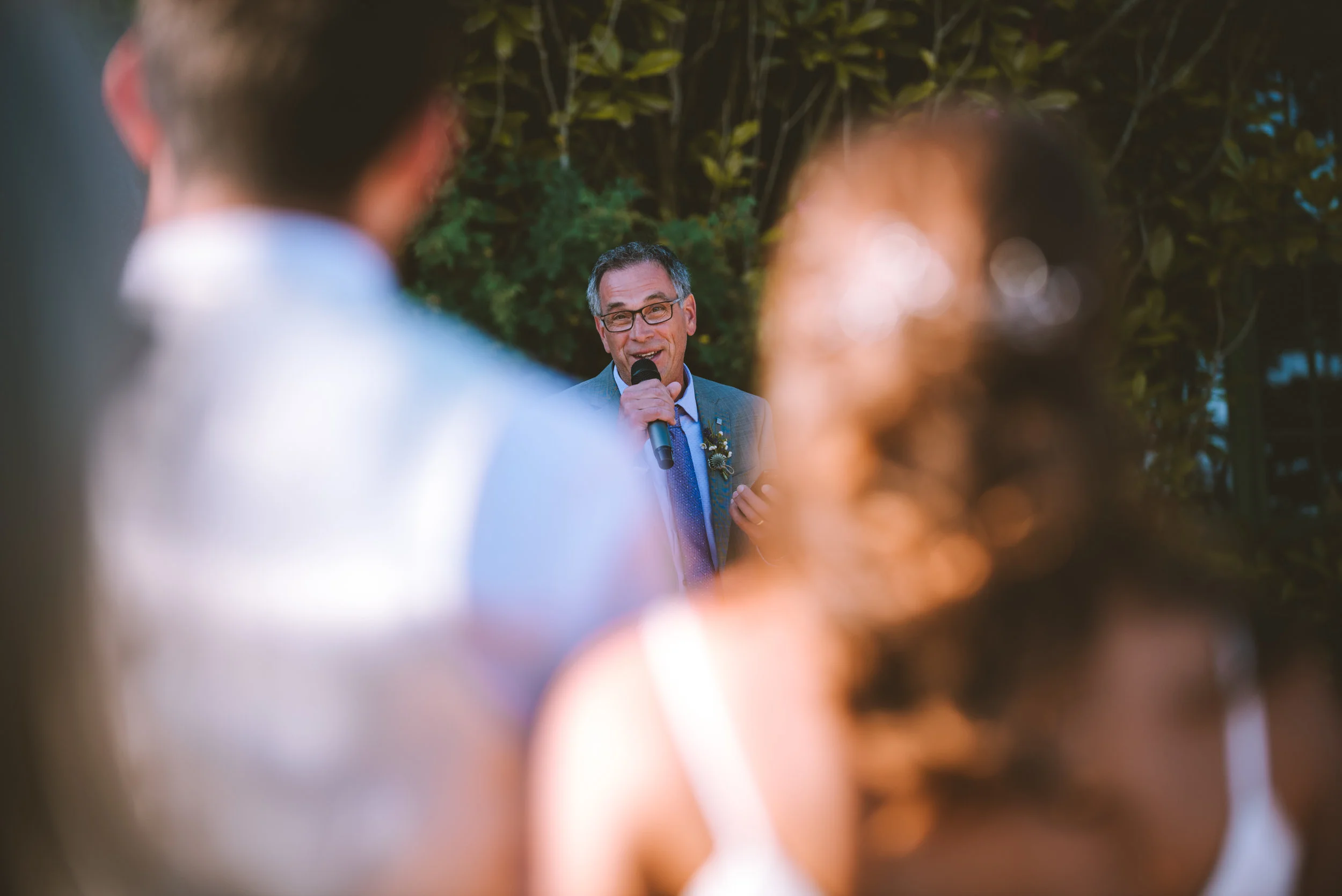 A man in glasses and a suit giving a speech at an outdoor event, with two blurred individuals in the foreground.