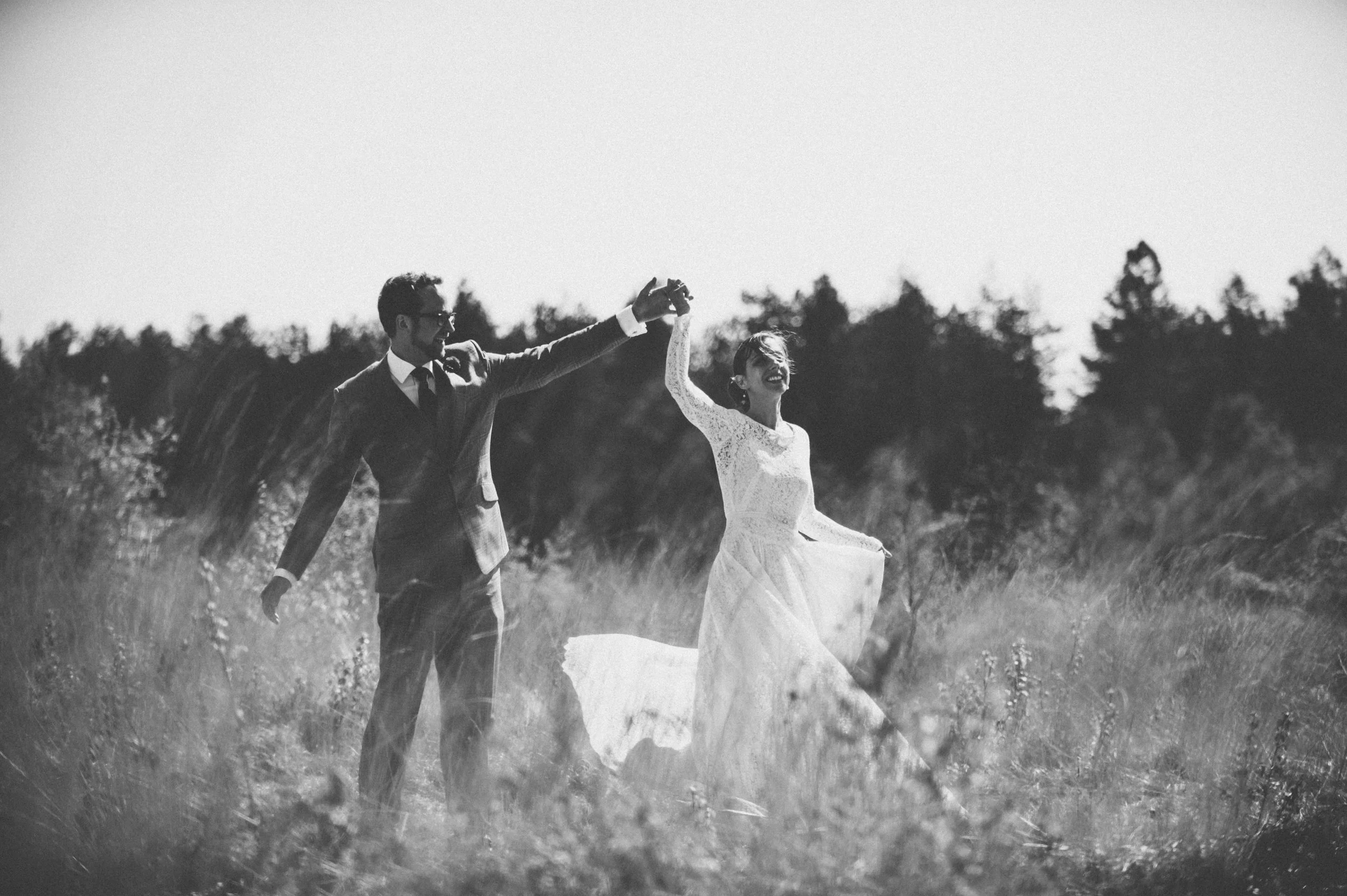 A black and white photo of a bride and groom outdoors, with the groom holding the bride's hand and twirling her as she smiles, in a grassy field with trees in the background.