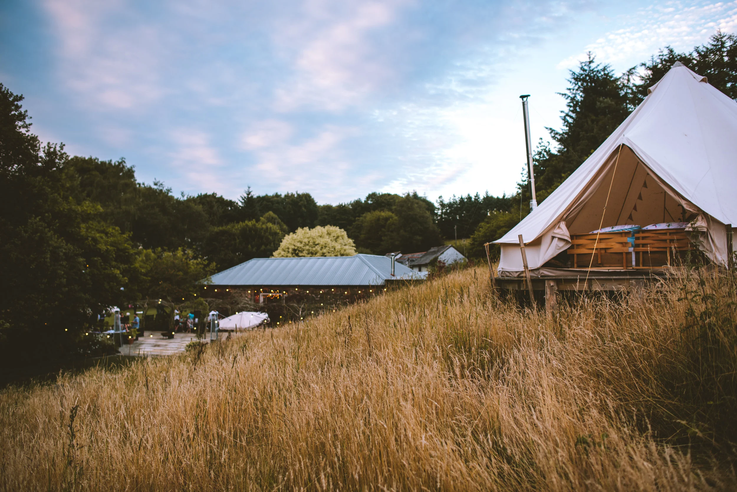A white canvas tent on a grassy hill with trees in the background and a building with string lights at sunset.