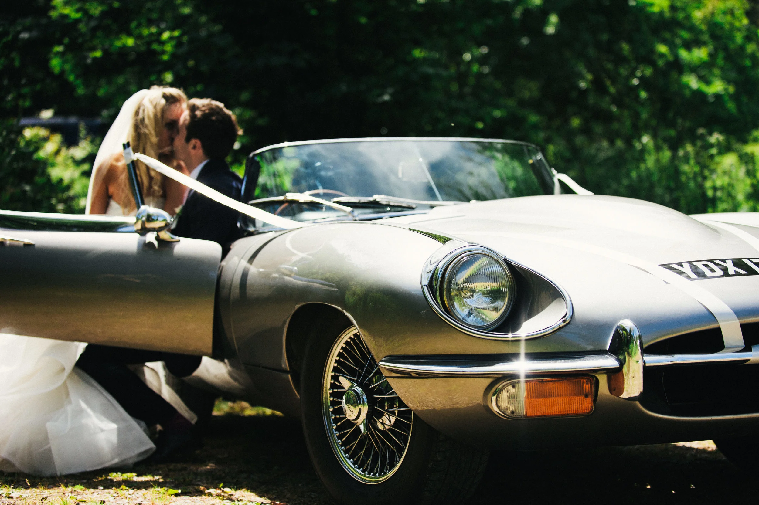 A bride and groom kissing in a vintage silver convertible car on a sunny day with green trees in the background.