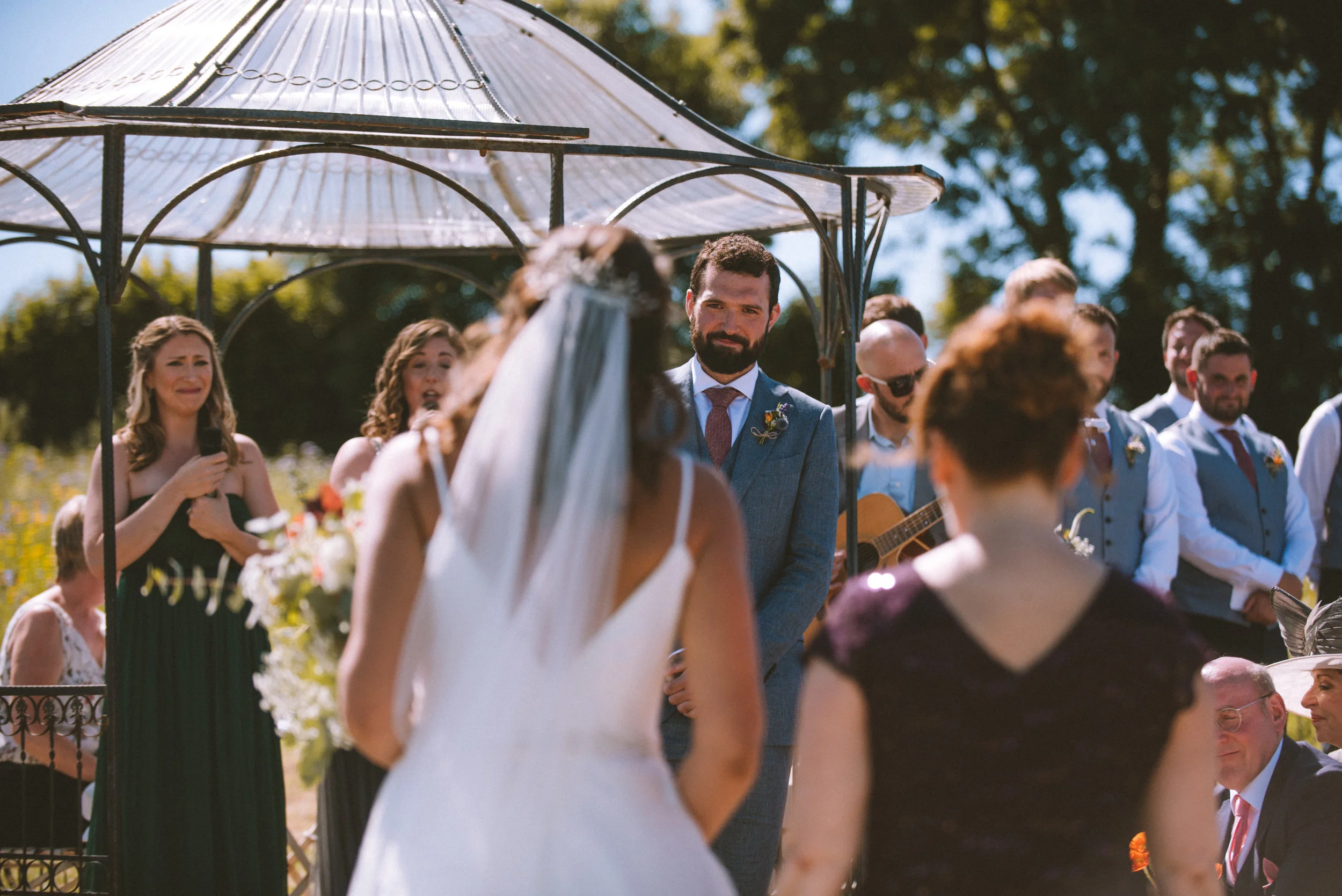 A wedding ceremony outdoors with the bride in a white dress and veil, standing in front of the groom in a gray suit, under a decorative metal gazebo with a domed roof. The bridal party and guests are visible, including a woman singing with a micropho