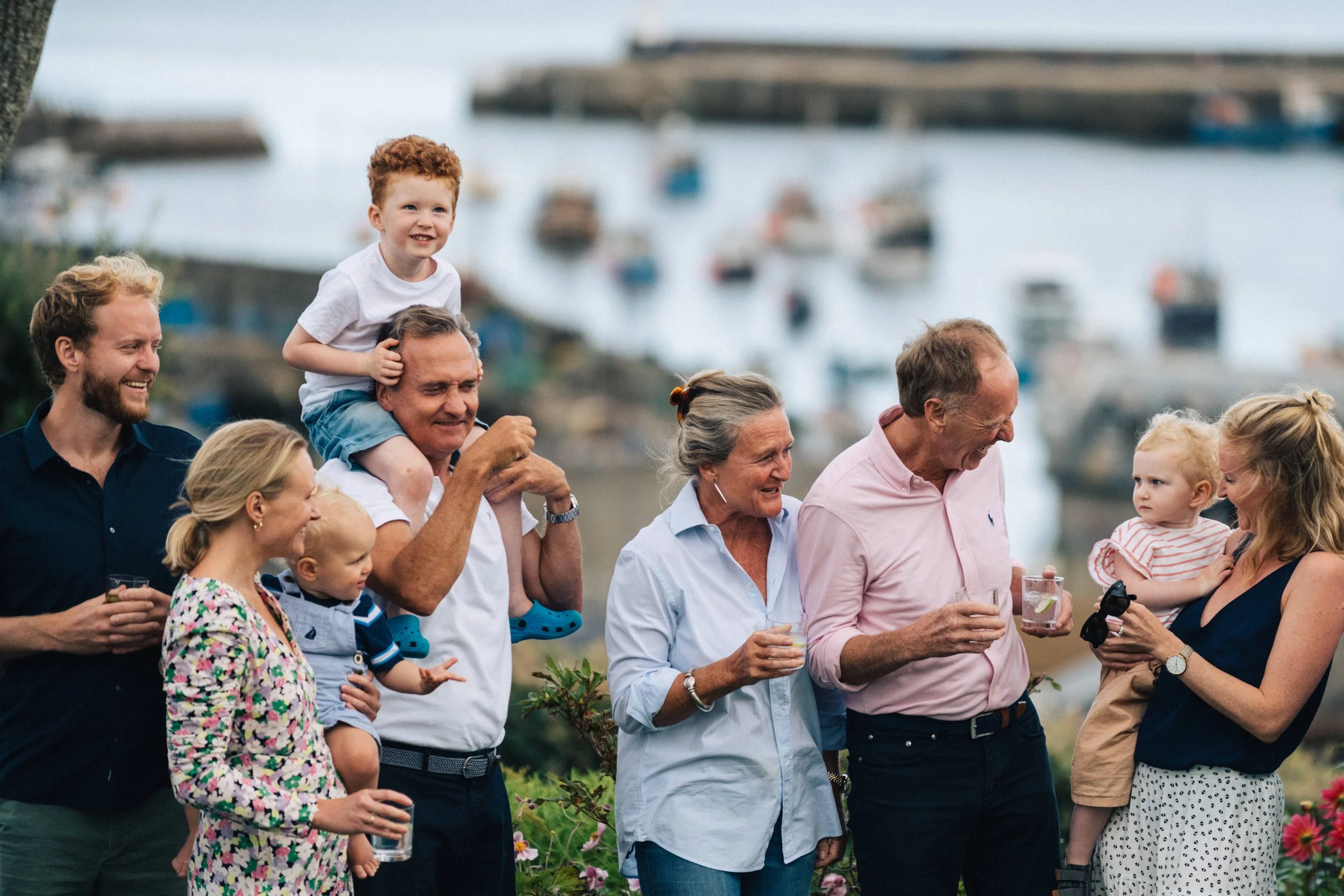 A group of eight people, including children and adults, gathered outdoors near a marina with boats. They are smiling and holding drinks, and one child is sitting on a man's shoulders.