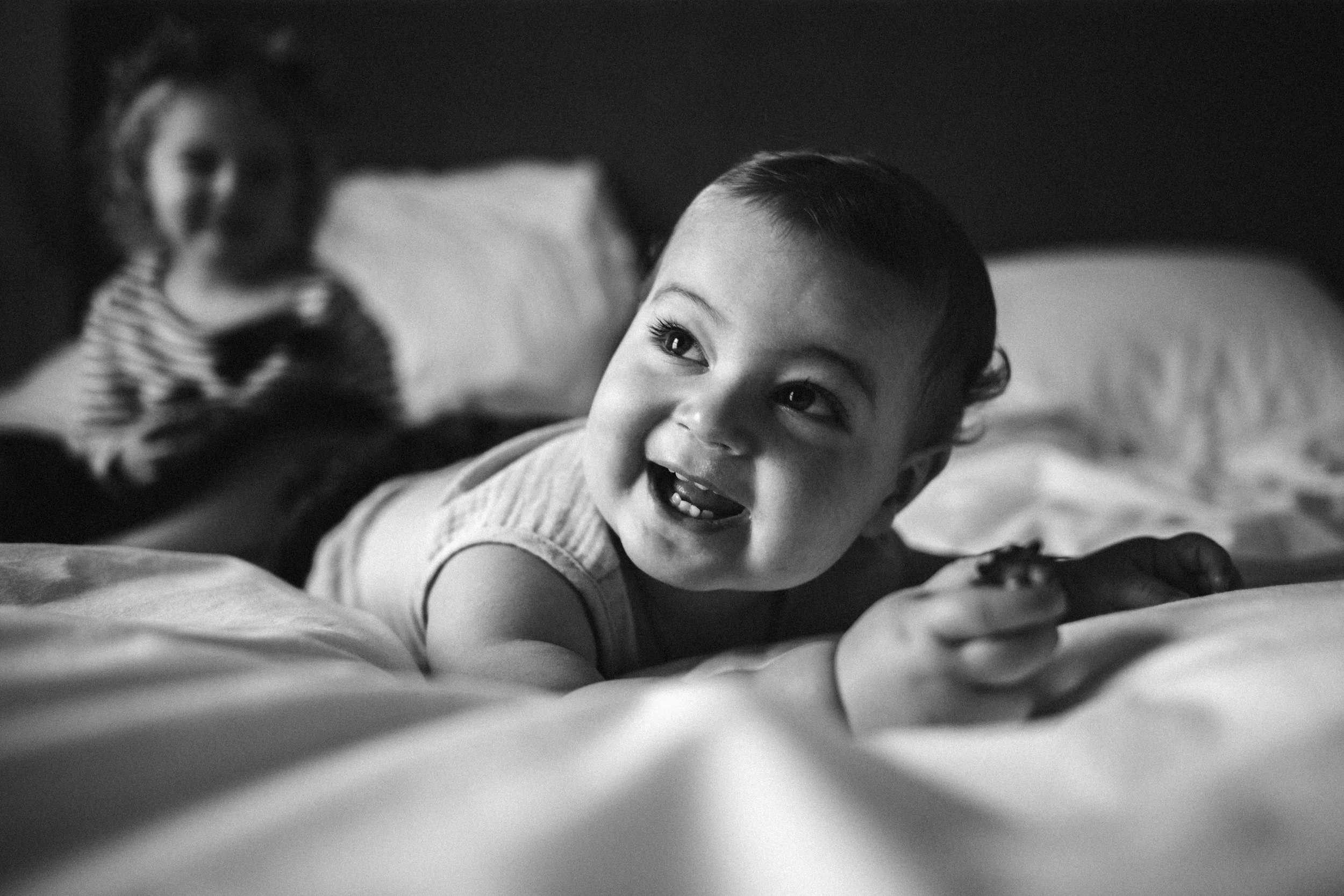 A young boy with short hair lying on a bed, smiling and looking to the side, with a child in the background partially out of focus.