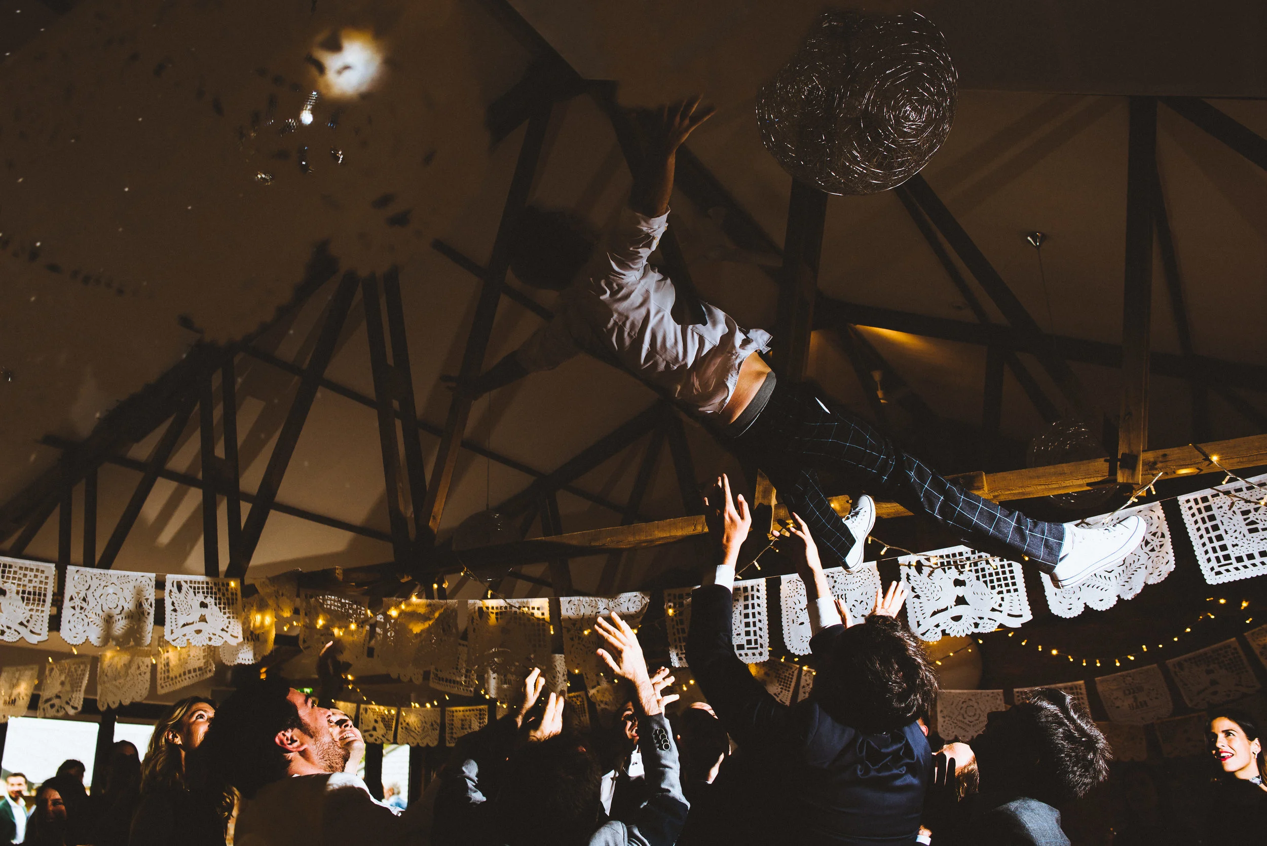 A person being hoisted into the air by a crowd at a celebration or event, with people reaching out. The setting is decorated with paper banners and string lights, in a dimly lit indoor space.