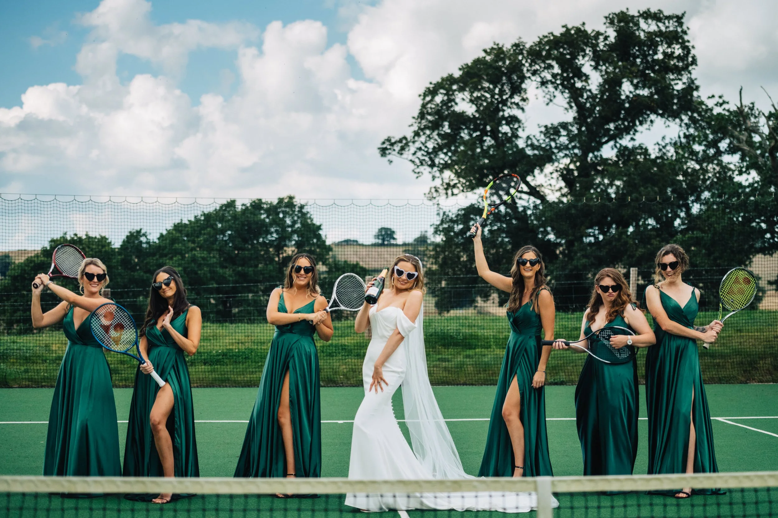 Bridal party on a tennis court, bride in white dress holding champagne, bridesmaids in green dresses holding tennis rackets, all wearing sunglasses.
