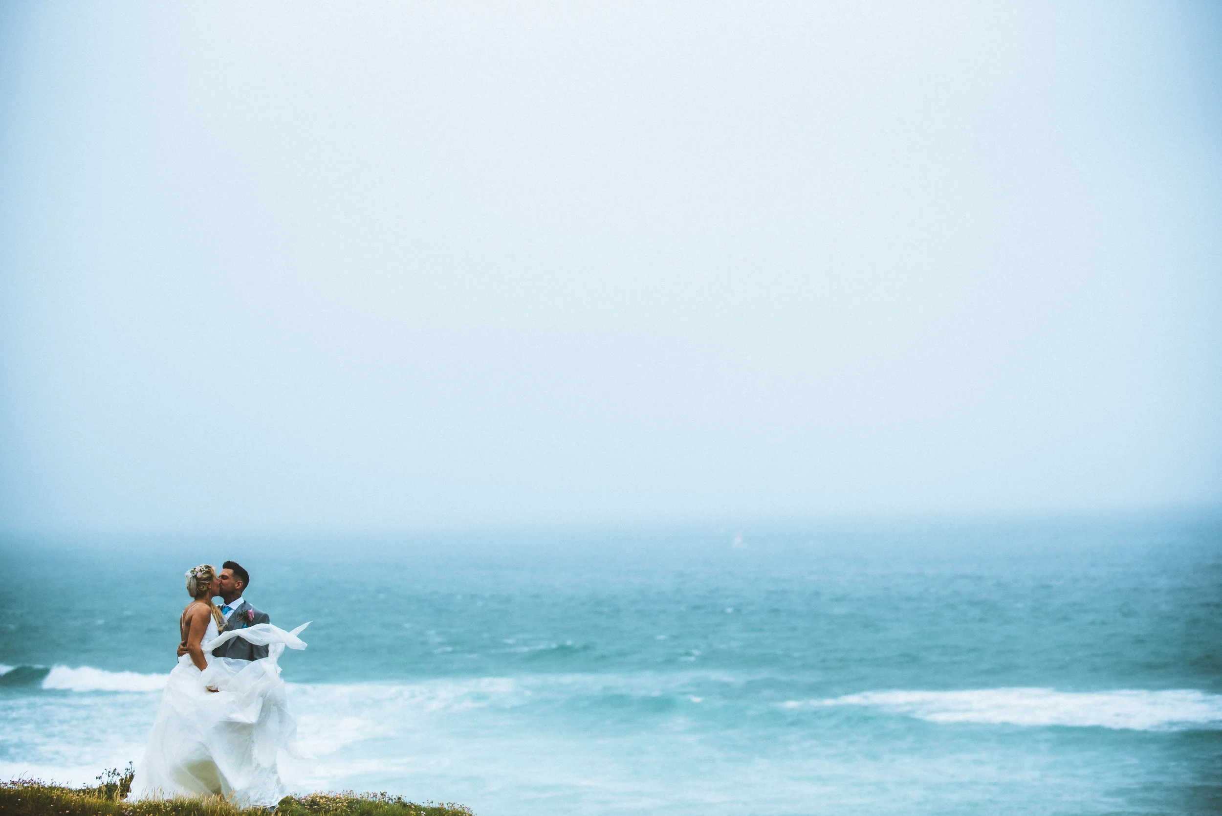Bride and groom romantic kiss on a Cornish coast in stormy weather, captured candidly by award-winning Cornwall wedding photographer, Mark Shaw. 