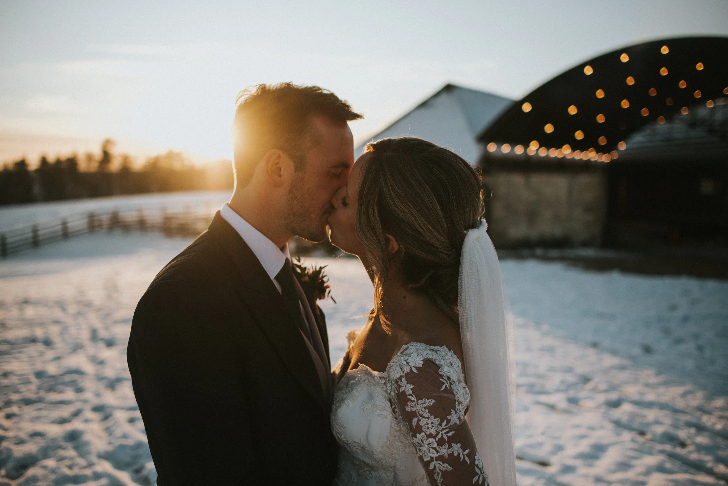 Bride and groom kissing in snowy outdoor setting at sunset with string lights in background.