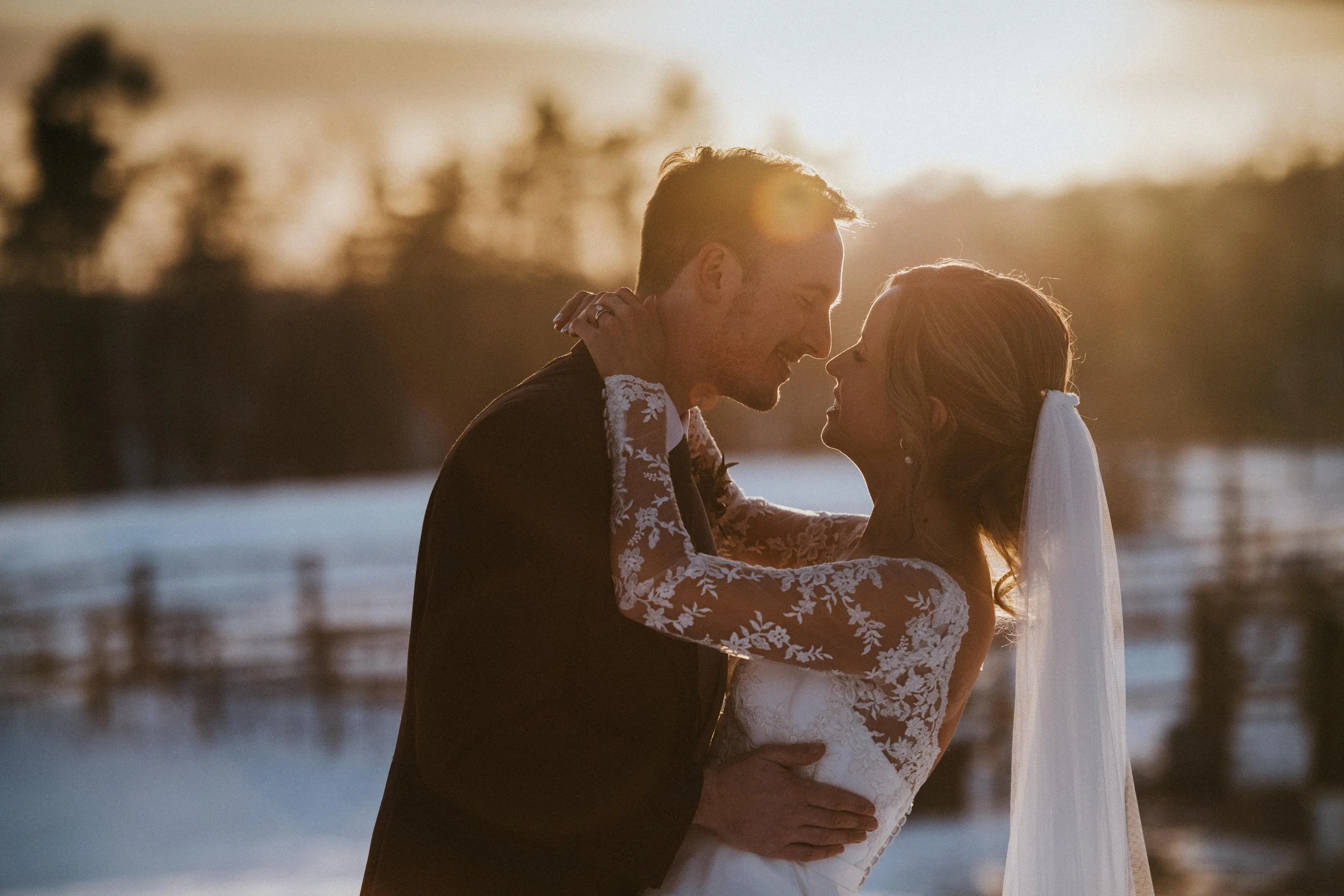 Bride and groom embracing at sunset, wearing wedding attire, with a blurred natural background.