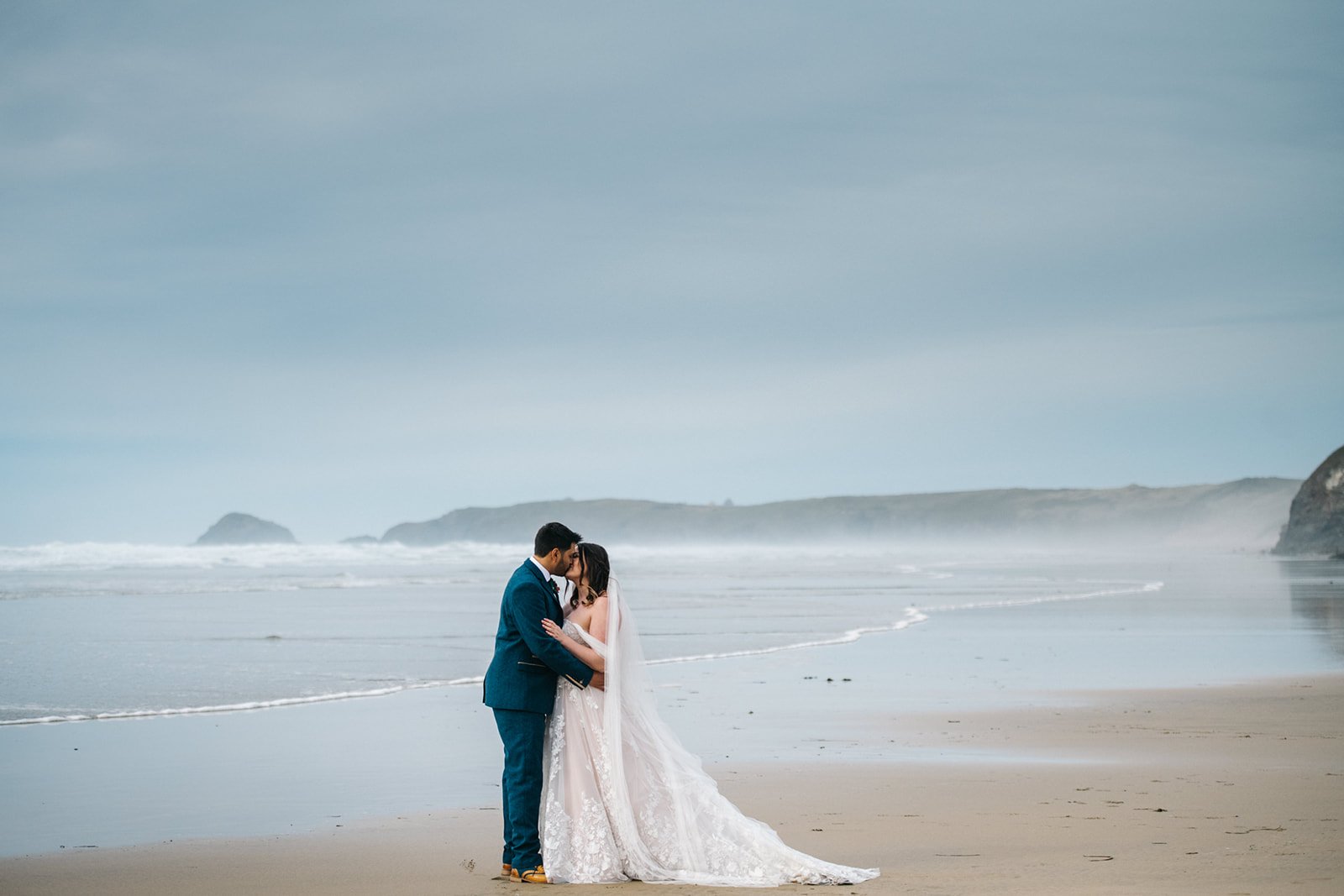 A couple dressed in wedding attire sharing a kiss on a beach with misty ocean waves and rocky cliffs in the background.