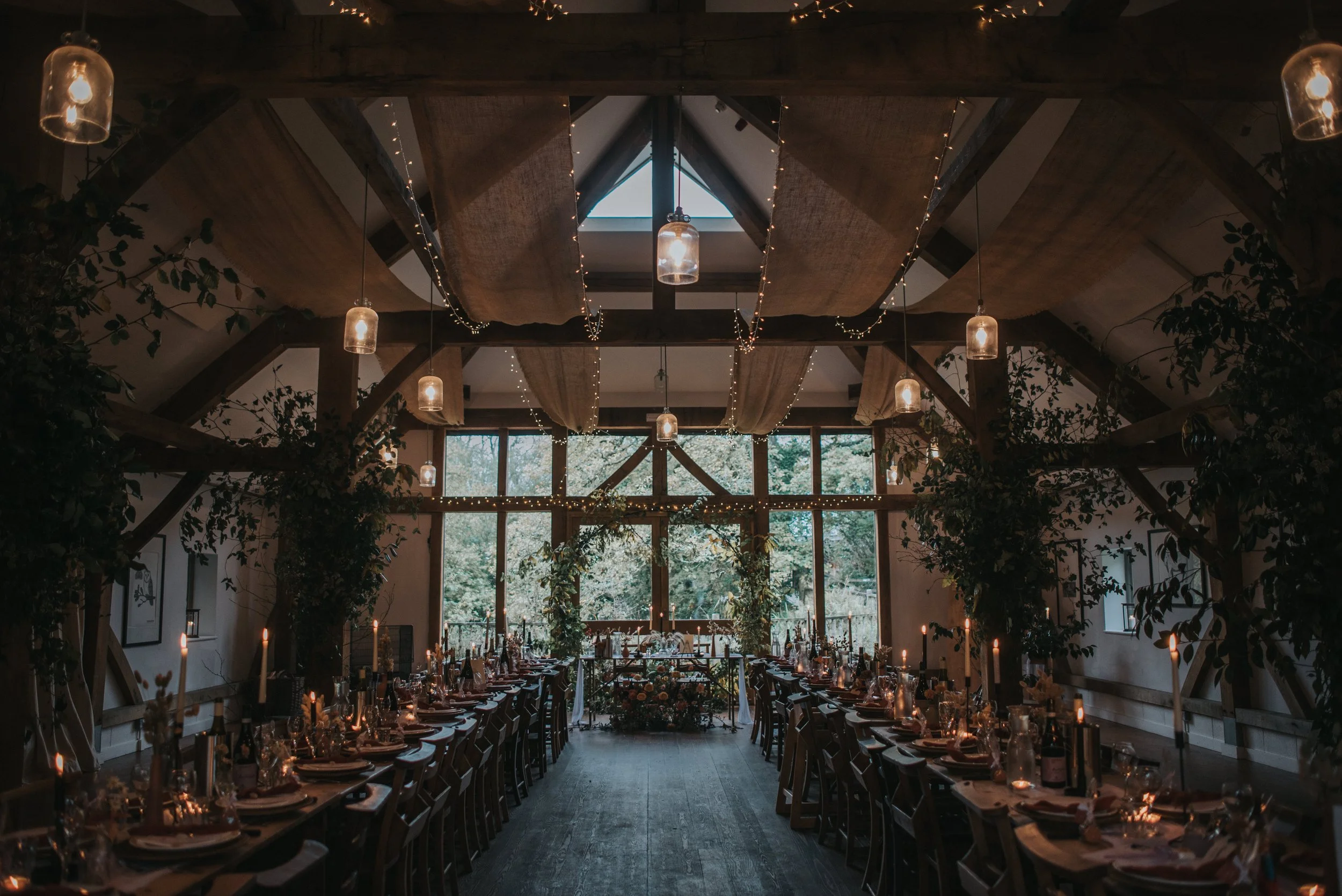 Rustic wedding breakfast setup from Nancarrow Farm in Cornwall with wooden beams, long dining tables, candles, hanging lights, greenery, and large windows.