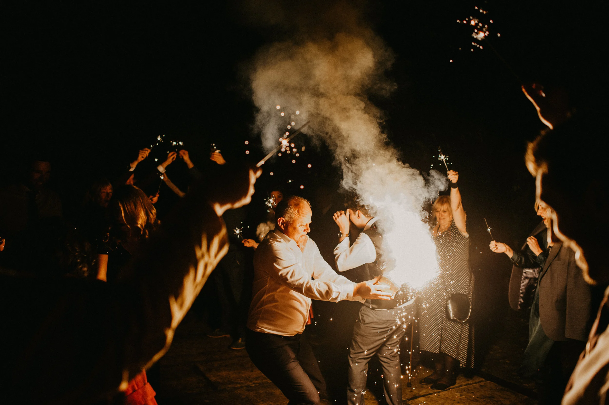 Wedding guests celebrating with sparklers at night, surrounded by smoke and darkness, captured by award-winning Cornwall wedding photographer, Mark Shaw.
