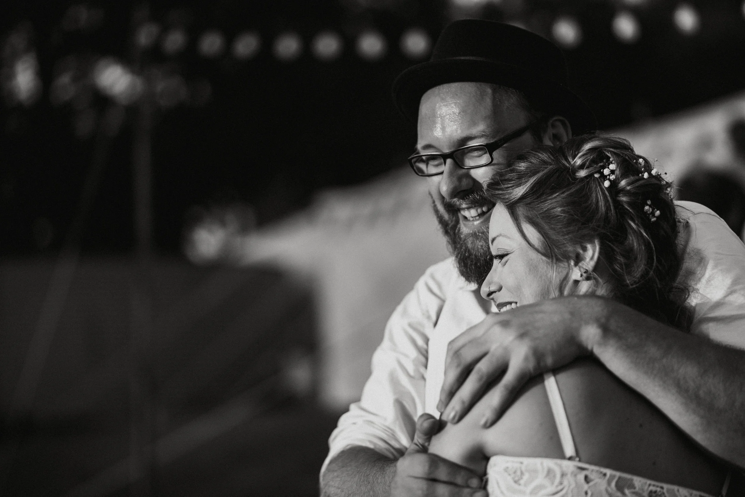 Black and white photo of a couple embracing and smiling, man wearing a hat and glasses, woman with styled hair and floral decorations, captured by award-winning Cornwall wedding photographer, Mark Shaw. 