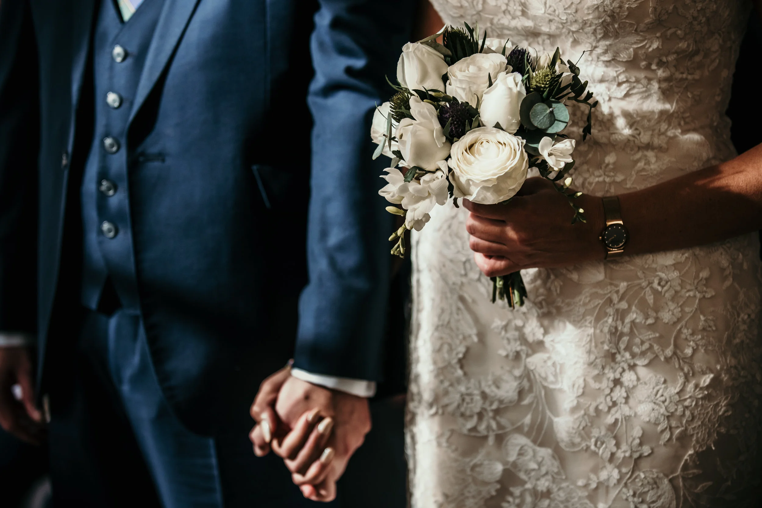 Wedding couple holding hands, bride in lace dress with bouquet, groom in blue suit.