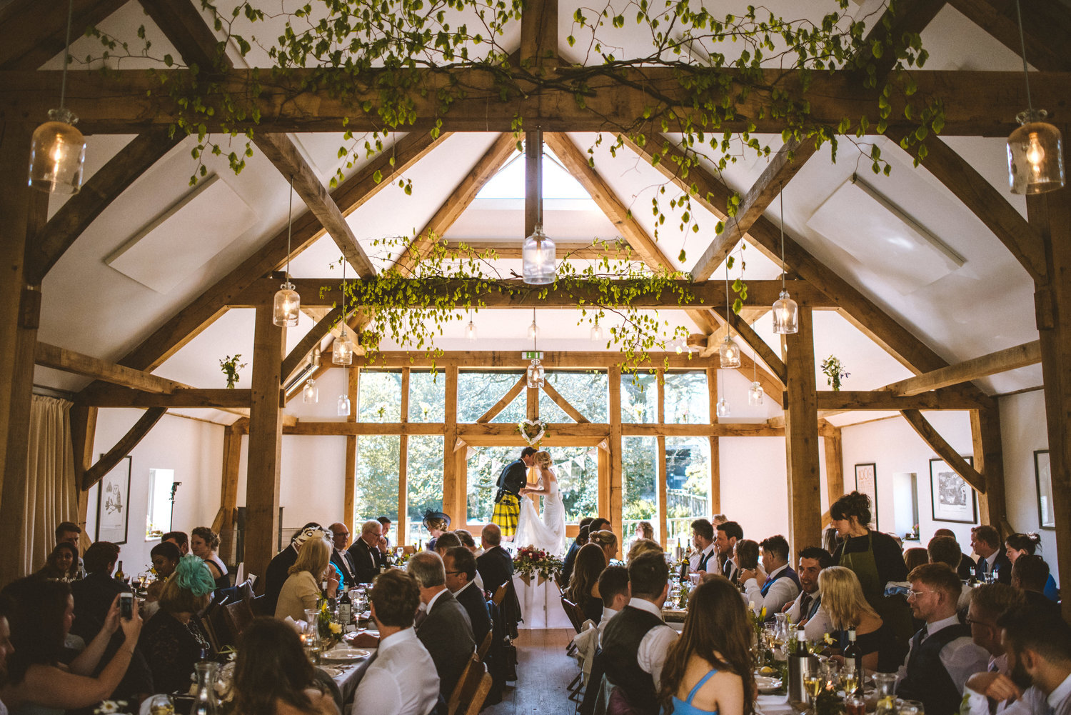 Rustic wedding reception in a barn with wooden beams and greenery, featuring a couple kissing while guests are seated at tables.