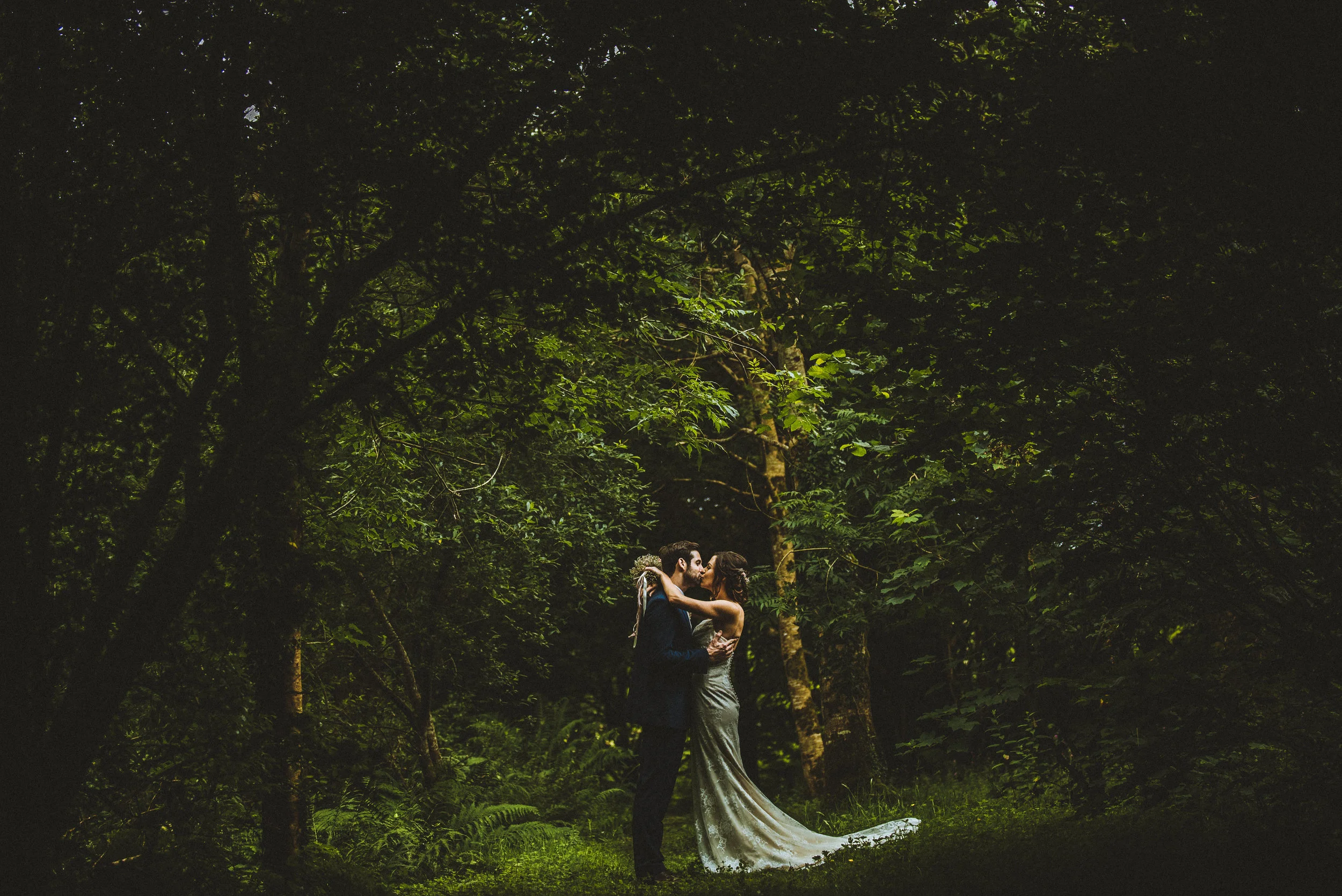 Couple kissing in a forest wearing formal attire, surrounded by lush greenery.