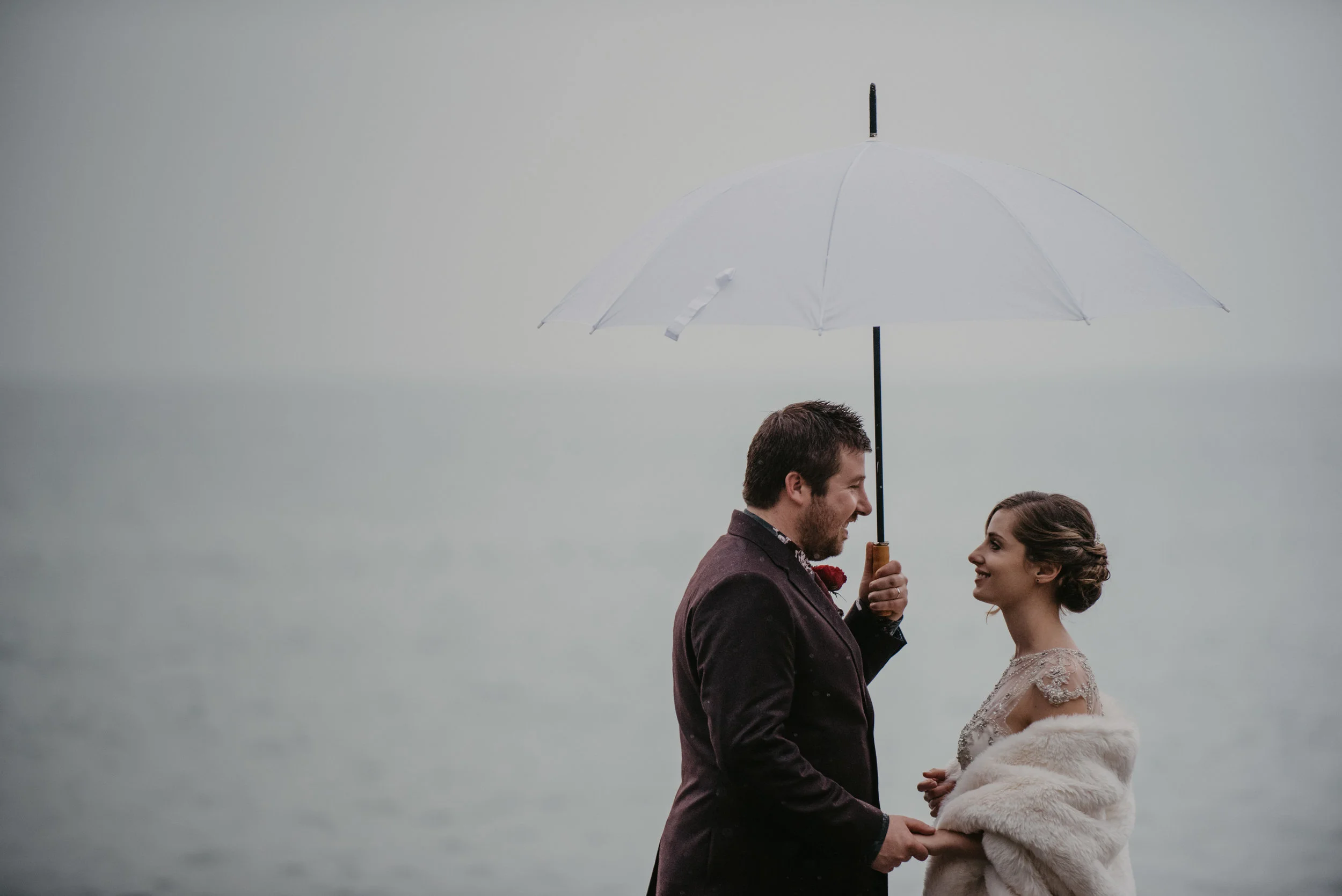 Couple standing under a white umbrella near a body of water, dressed in formal attire.