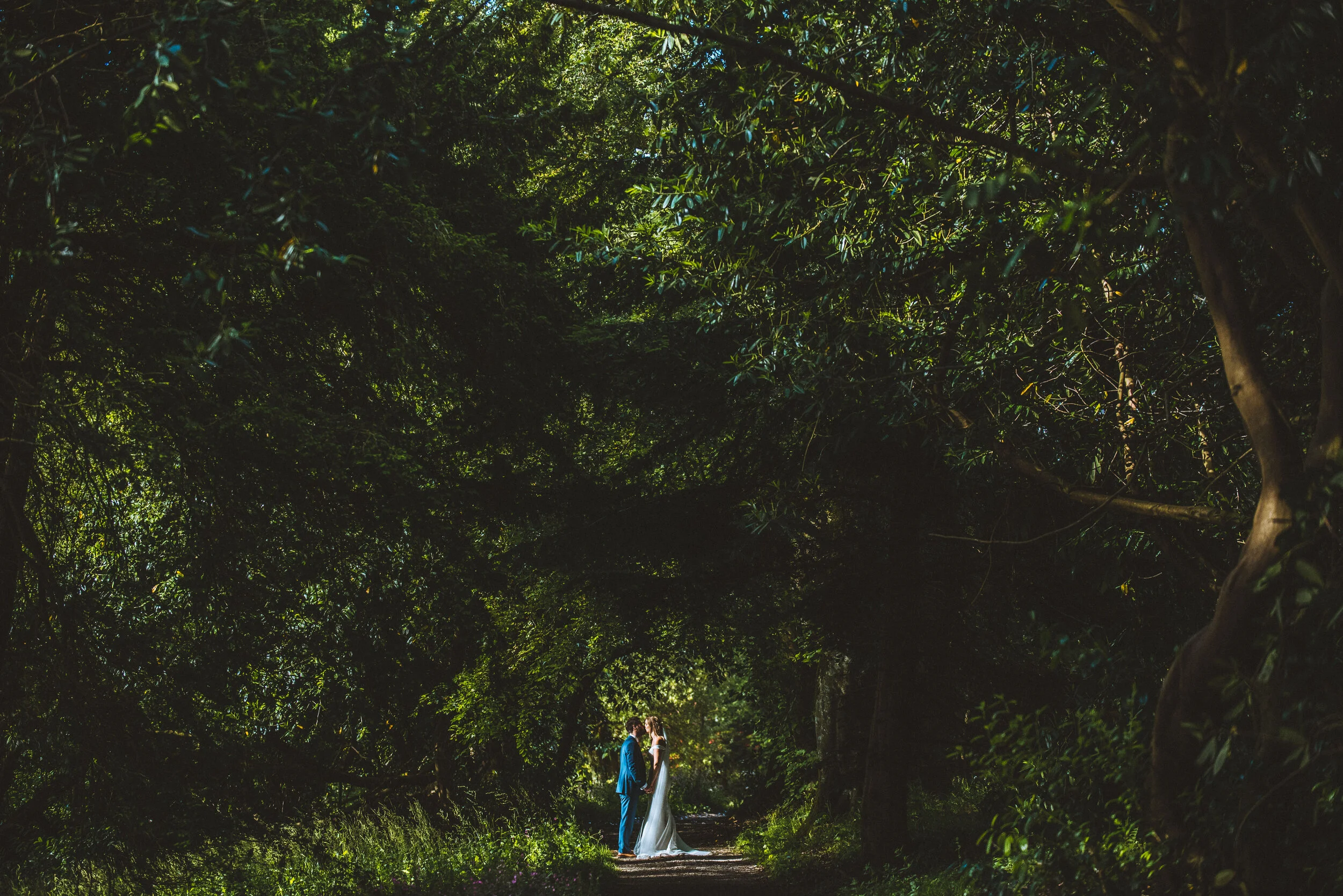 Bride and groom facing each other surrounded by lush green trees in the beautiful gardens at Pencarrow House, captured by award-winning Cornwall wedding photographer, Mark Shaw. 