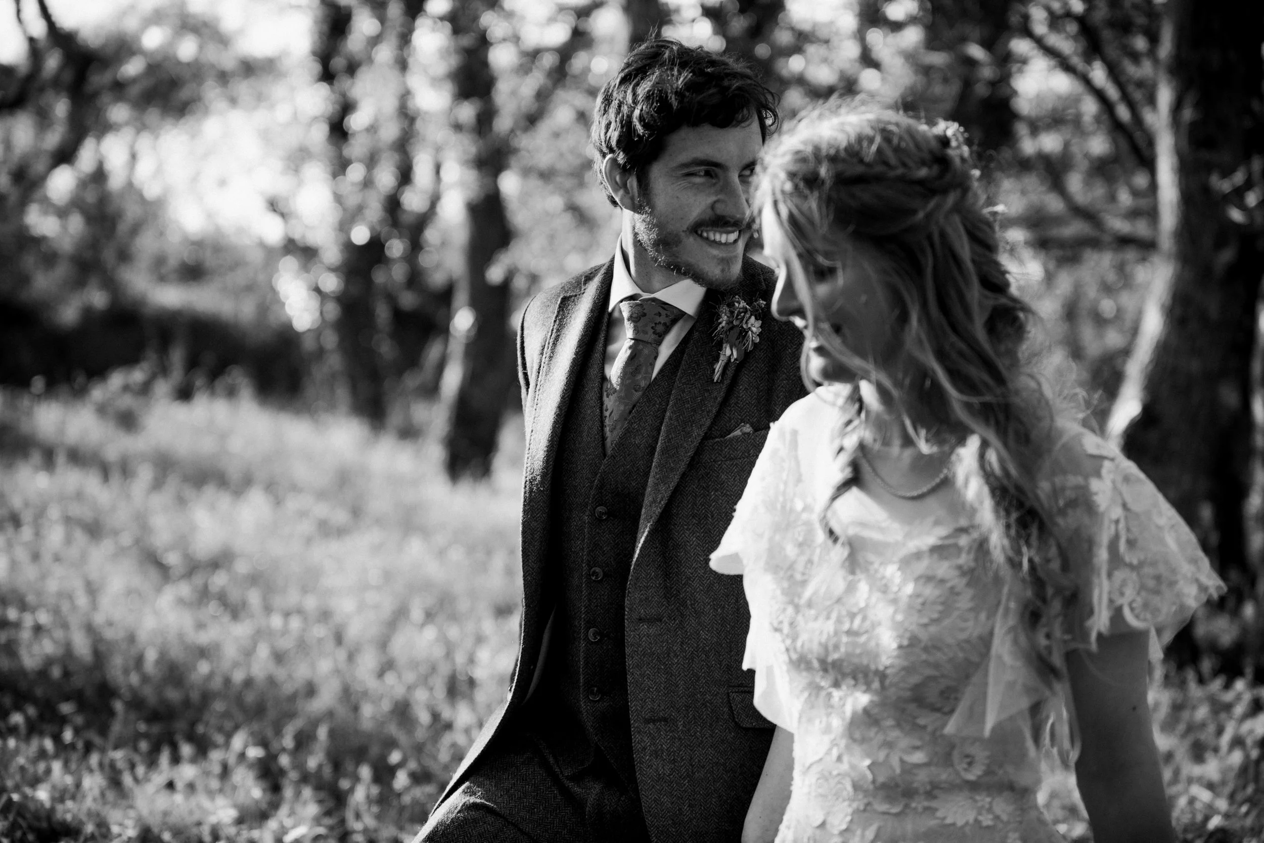 Black and white photo of a smiling couple in wedding attire walking outdoors, surrounded by trees, captured by award-winning Cornwall wedding photographer, Mark Shaw. 