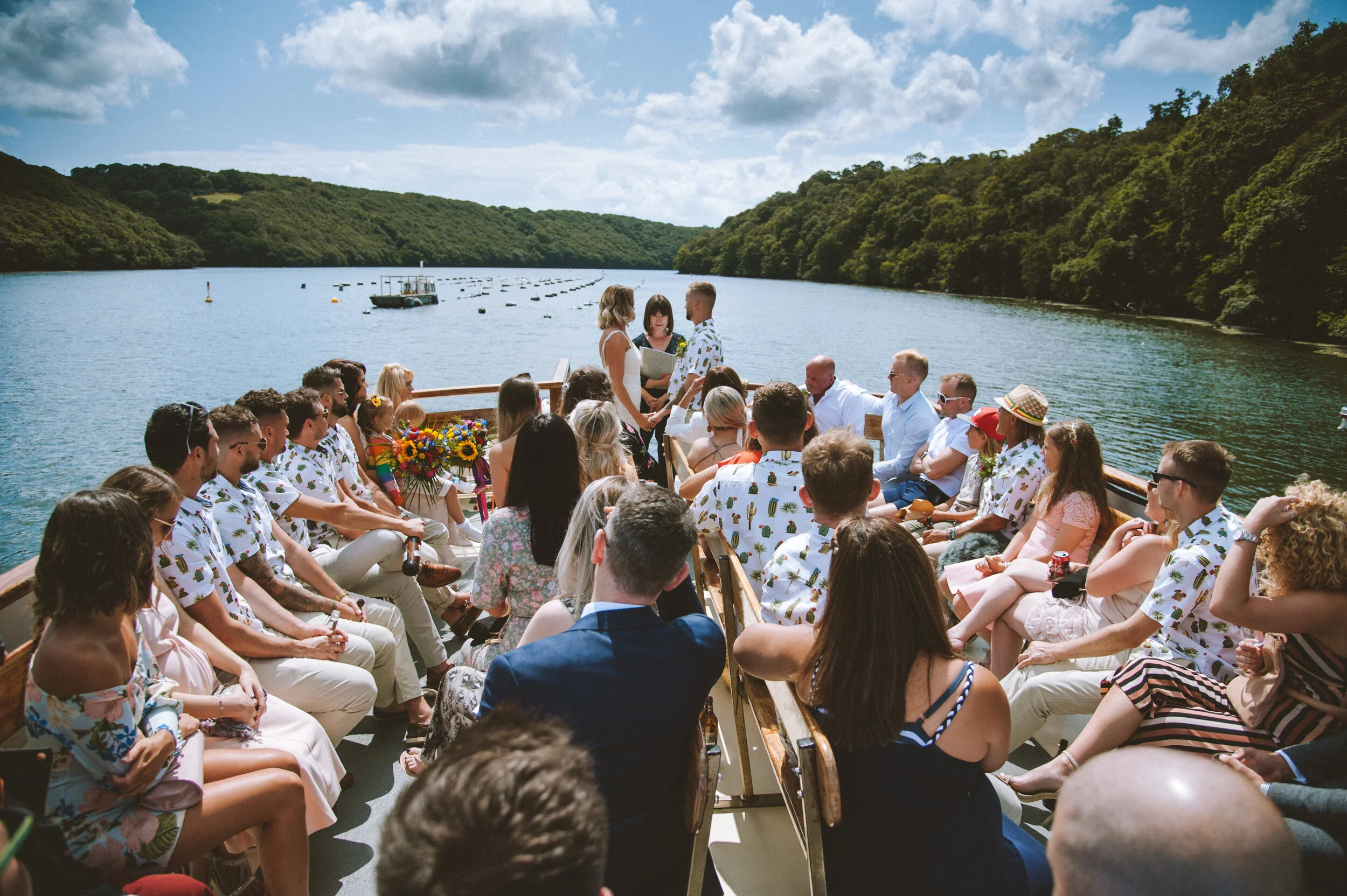 Outdoor boat wedding ceremony with guests, lake and hills in the background on a sunny day.