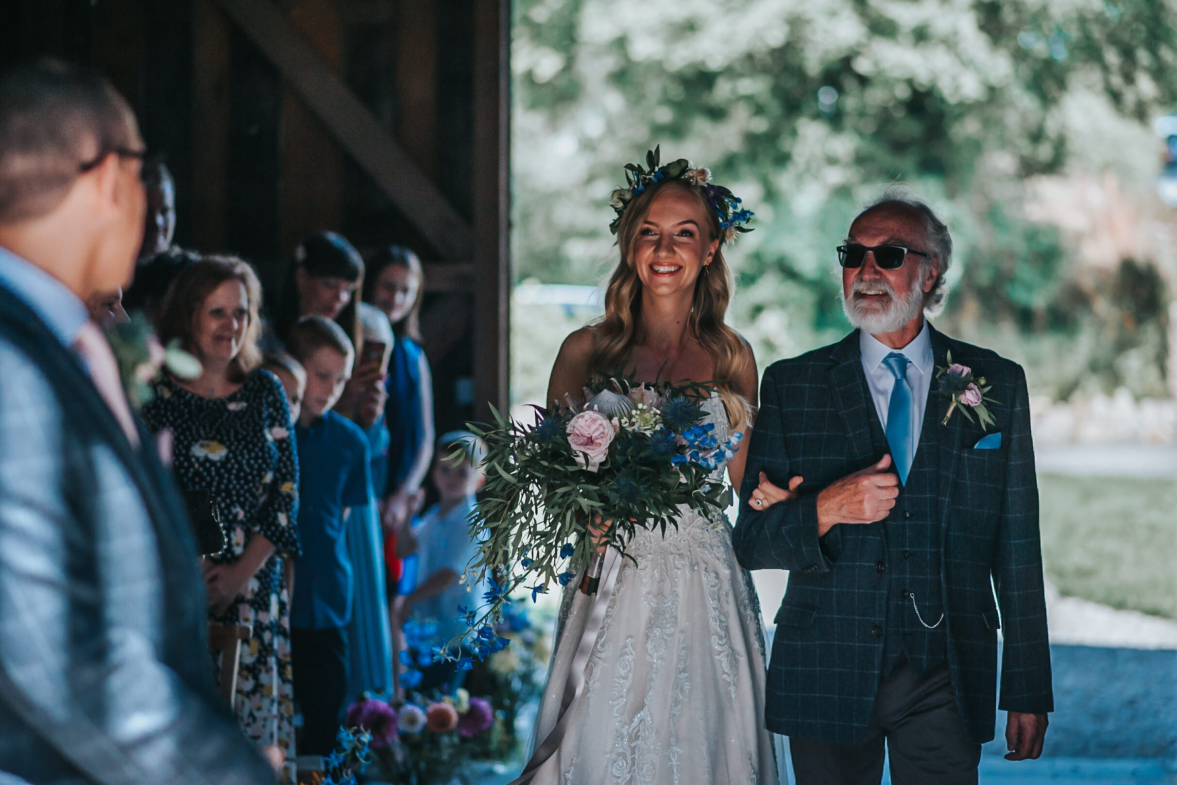 Bride with floral crown walking down the aisle at Nancarrow Farm with her dad, captured by award-winning Cornwall wedding photographer, Mark Shaw. 
