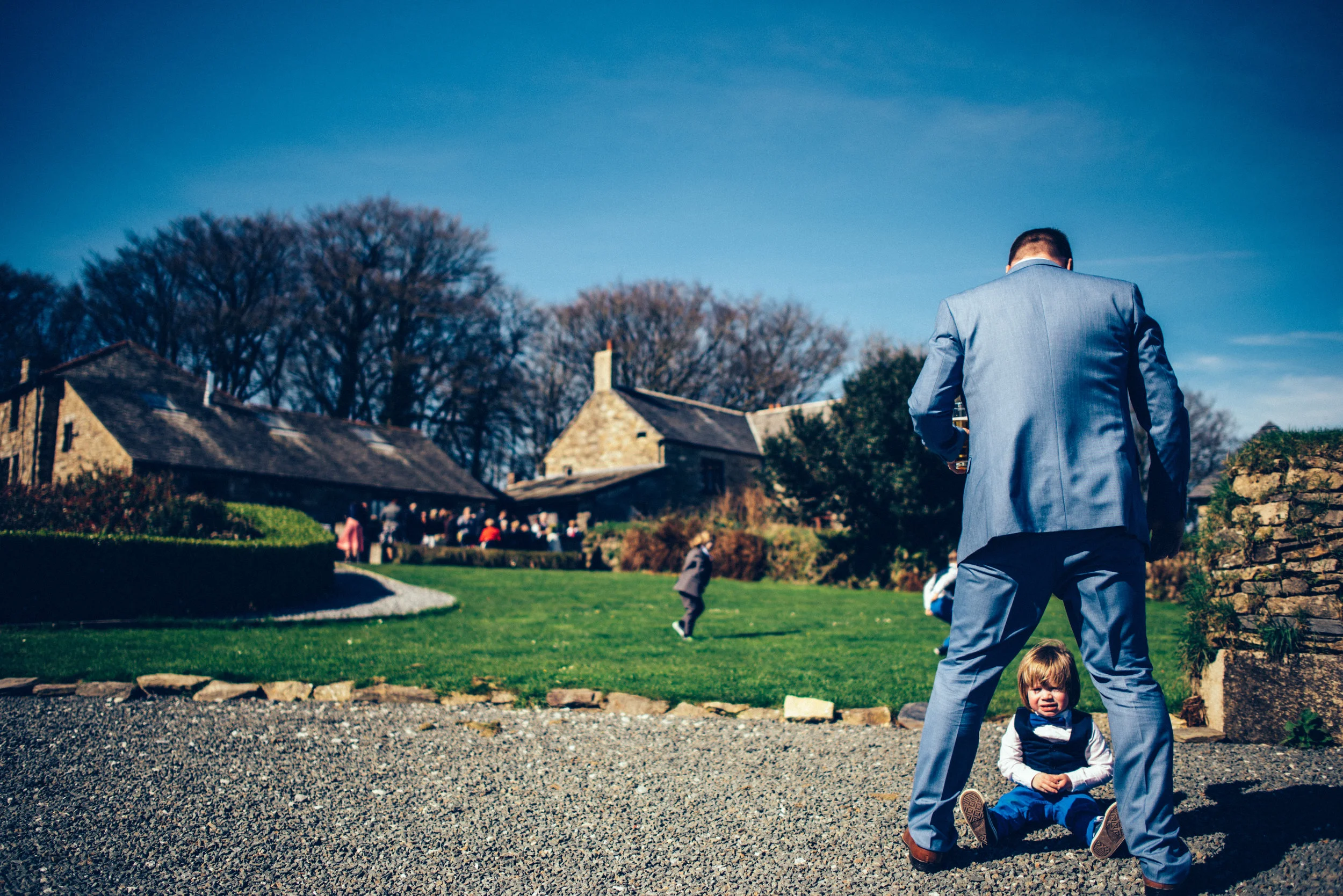 A man in a blue suit stands with his back to the camera, shielding a young girl who is sitting on the ground and looking at the camera in front of him. The scene is outdoors in a park or garden with green grass, leafless trees, and a stone building, 
