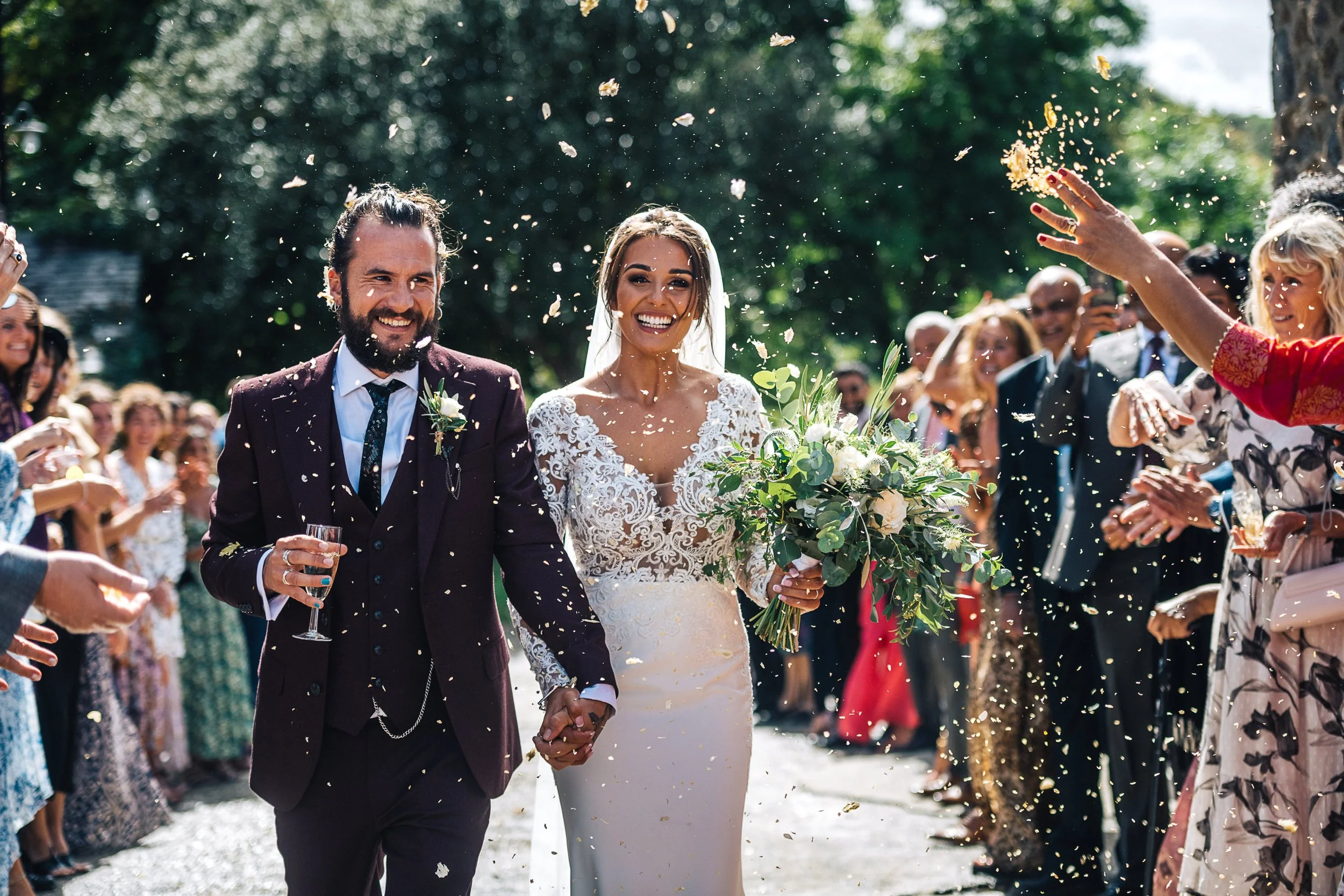 Bride and groom walking through confetti, surrounded by guests celebrating outdoors.