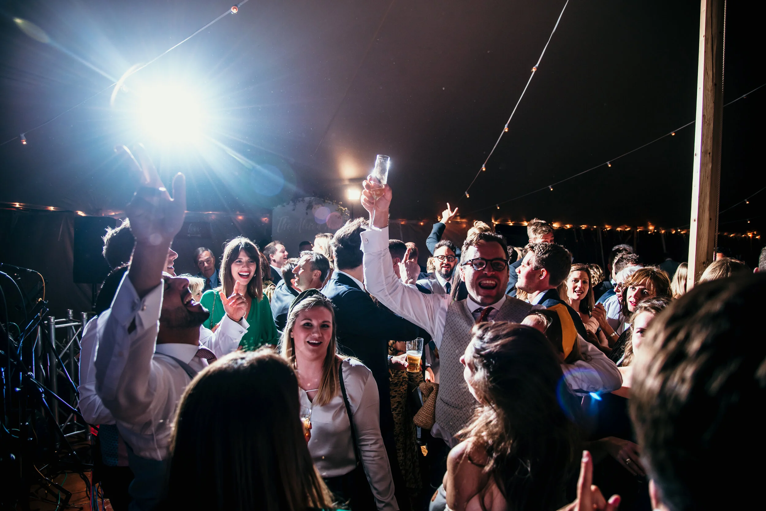 Wedding guests celebrating at a marquee party in Cornwall with drinks in hand, under string lights.