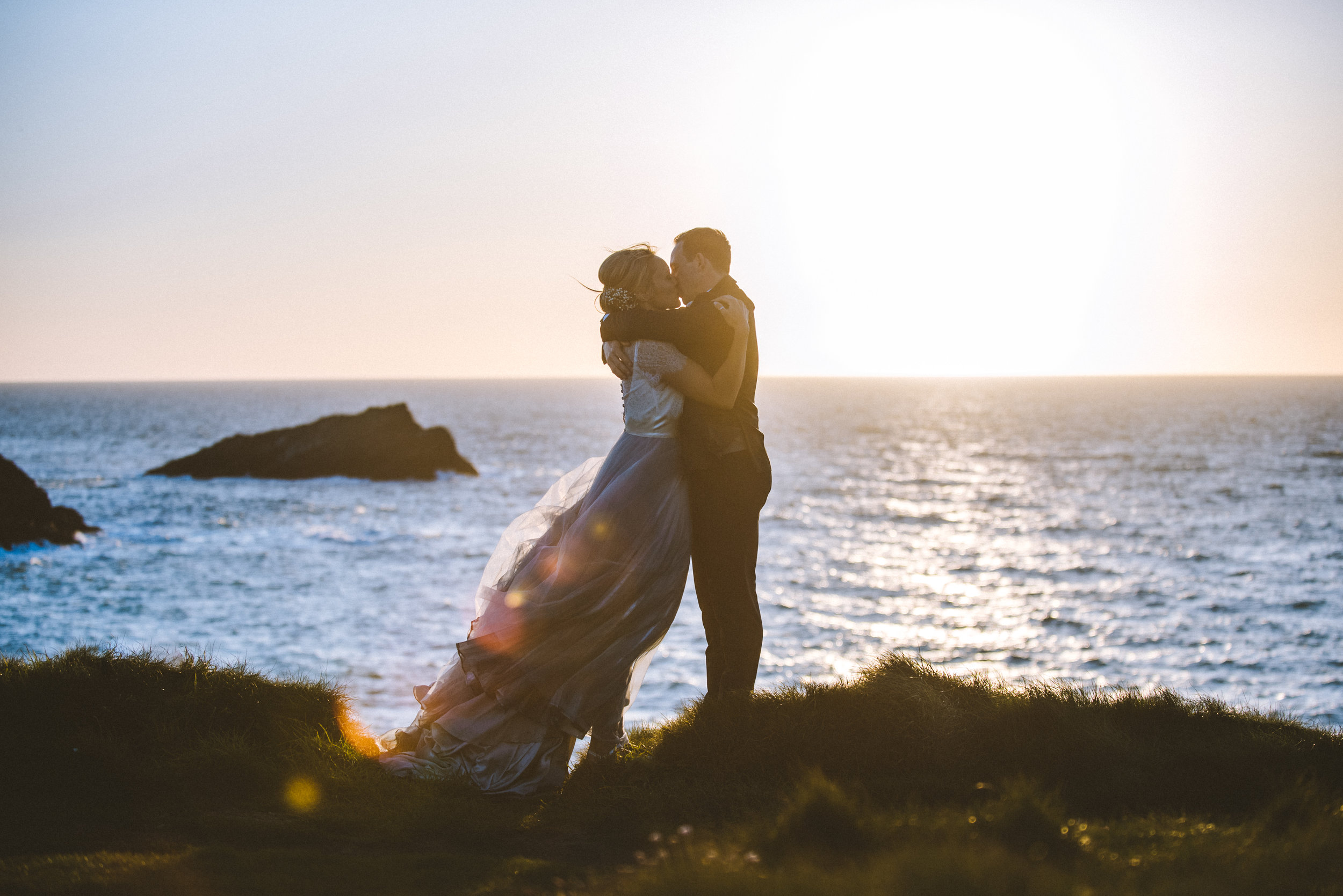 Couple embracing by the sea at sunset, with waves in the background, captured by award-winning Cornwall wedding photographer, Mark Shaw. 