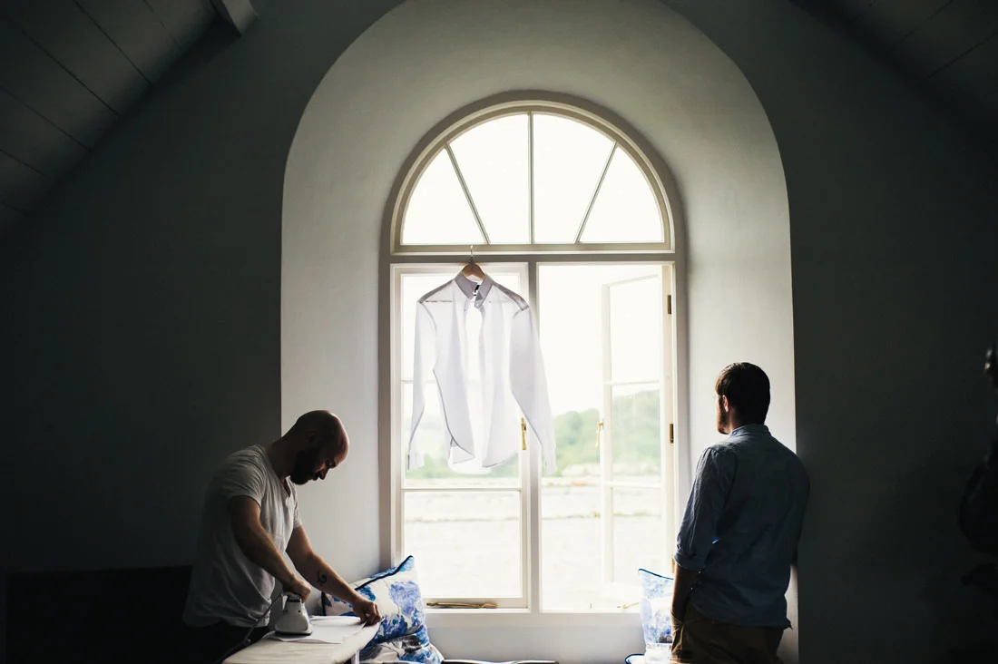 Two men in a dimly lit room, one ironing a shirt, the other looking out an arched window where a shirt hangs.