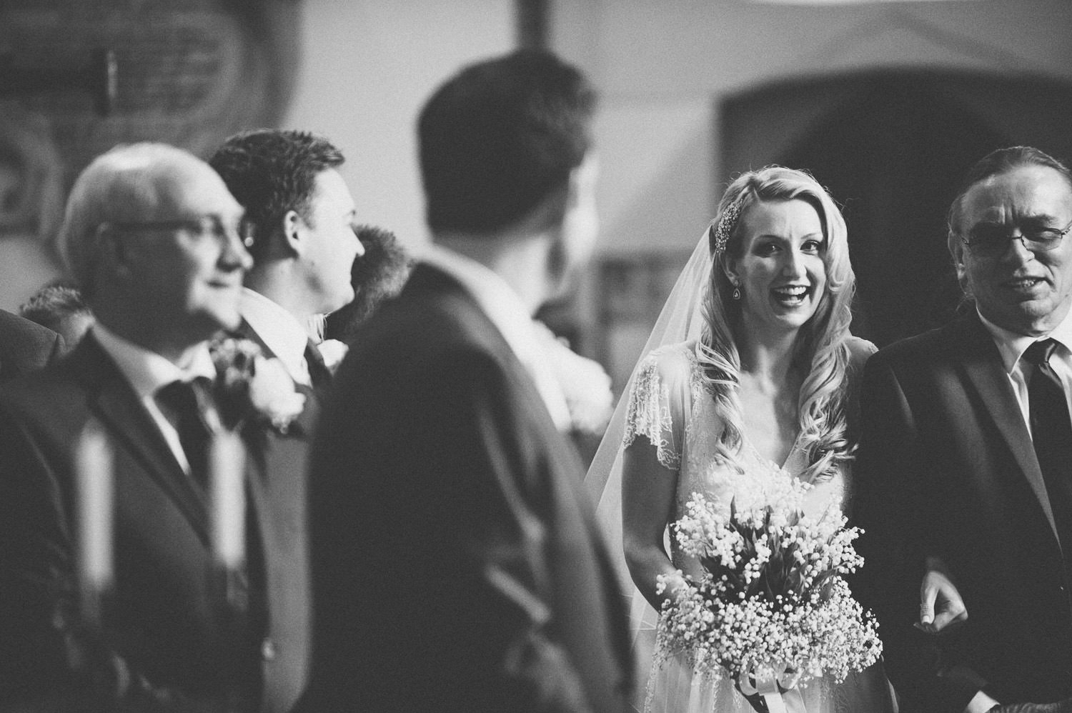 Black and white photo of a wedding ceremony, with a bride holding a bouquet of flowers smiling as she walks down the aisle, surrounded by several men in suits.