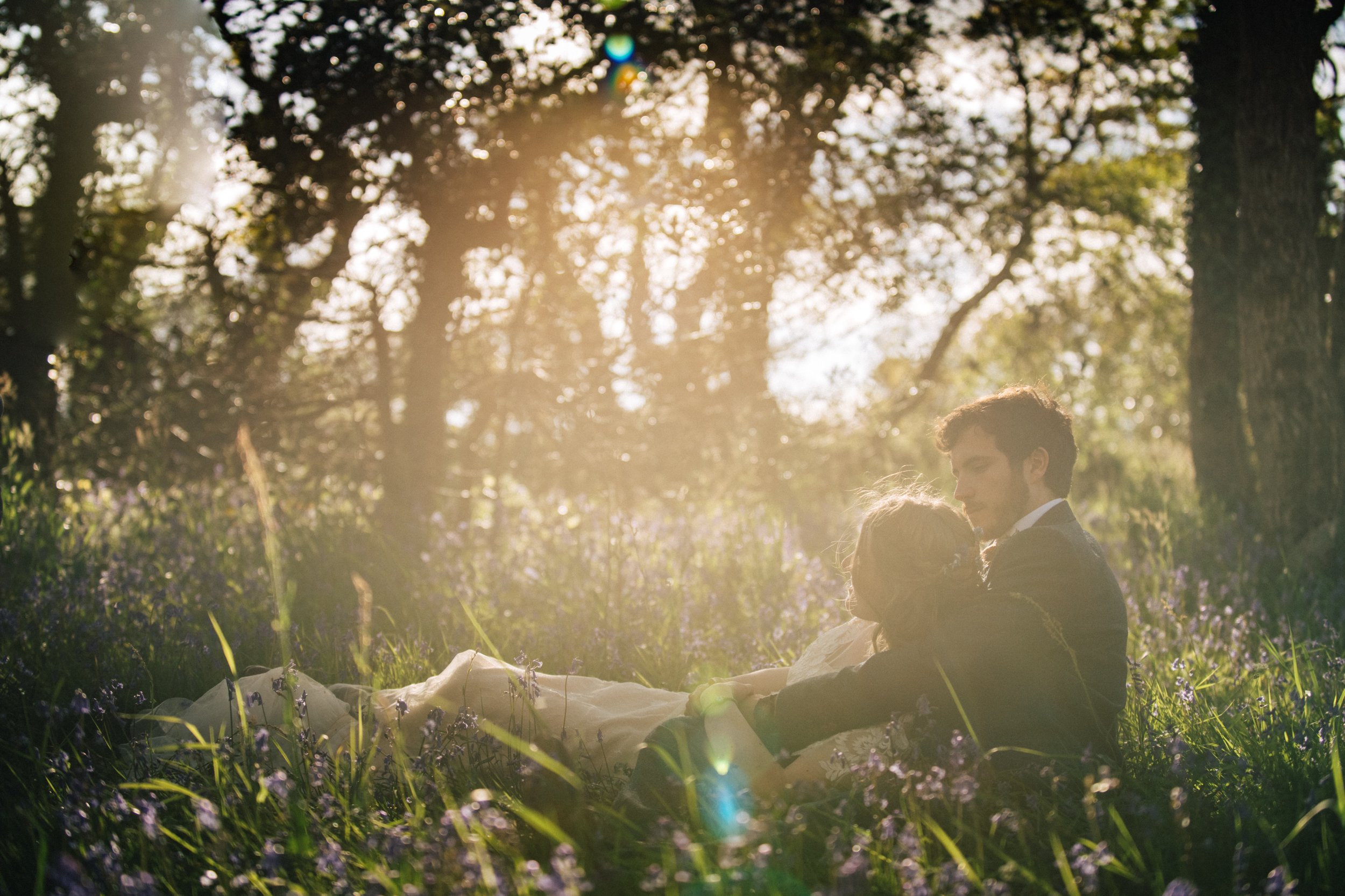 Couple sitting together in a sunlit forest clearing with flowers.