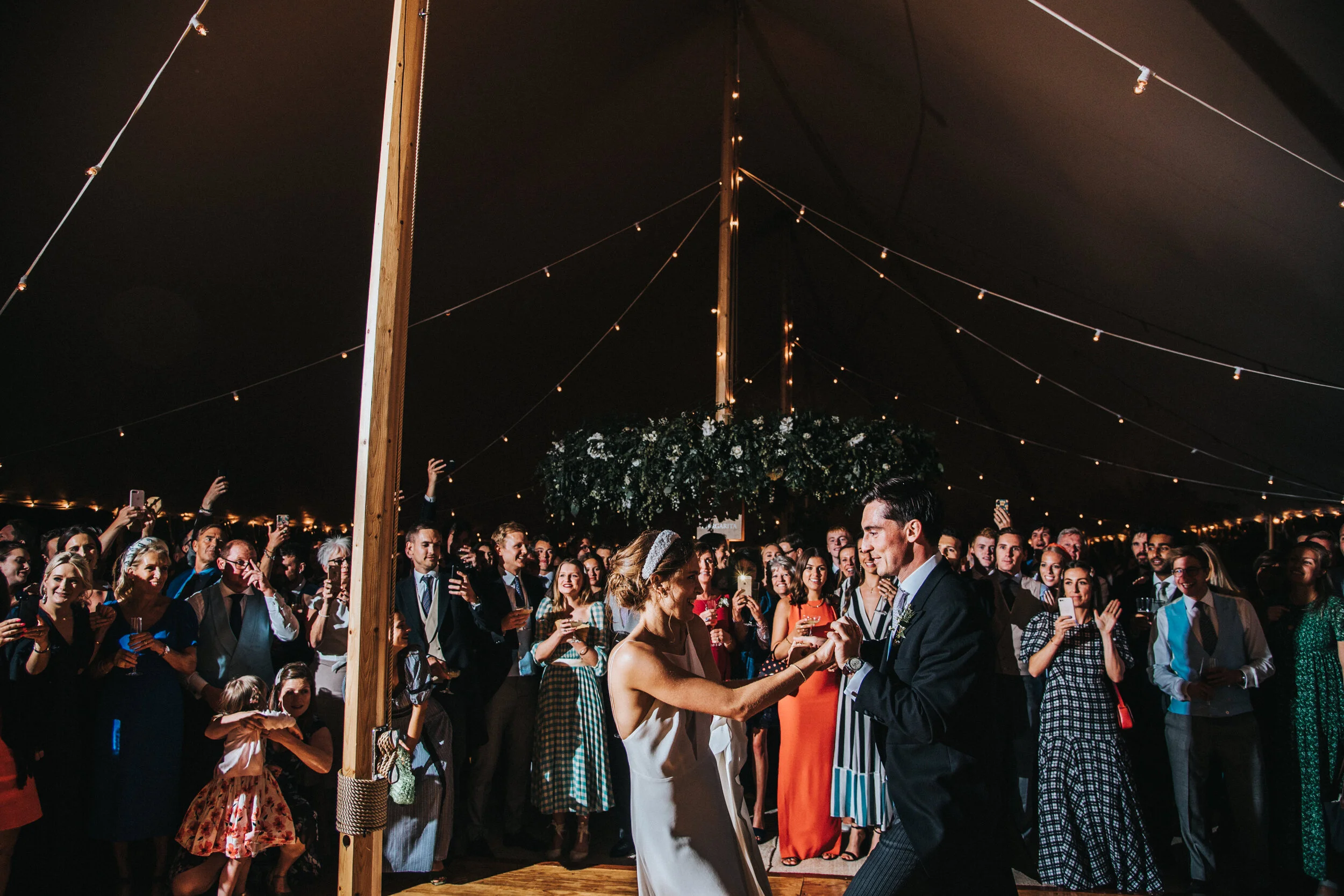 A couples first dance at a marquee wedding reception under a tent with string lights, surrounded by an audience, captured by award-winning Cornwall wedding photographer, Mark Shaw. 