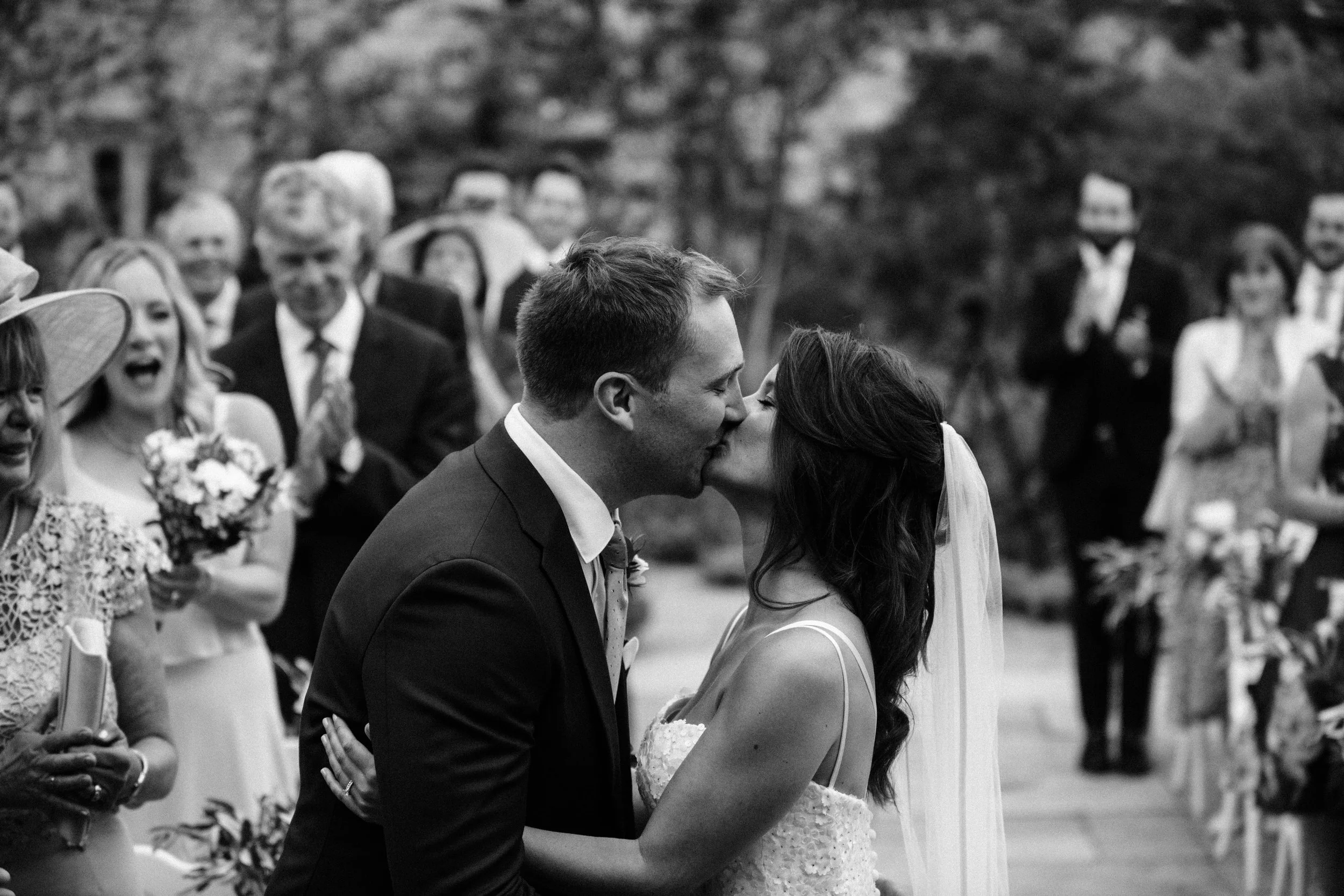 Bride and groom kiss during wedding ceremony with guests clapping and celebrating in the background.