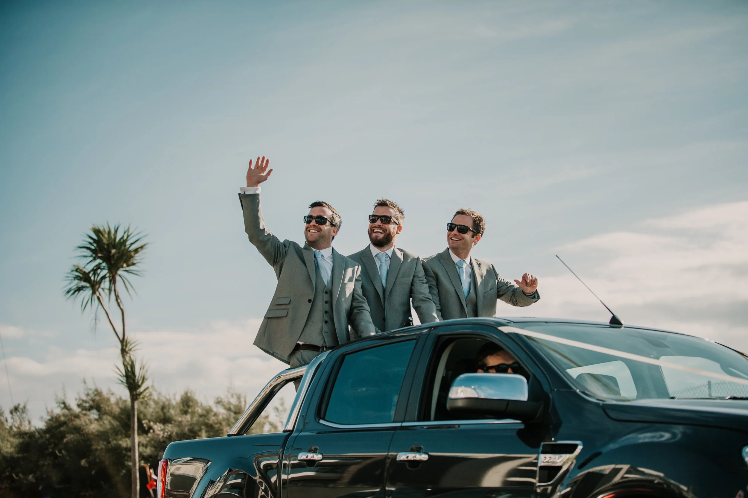 Three men in suits and sunglasses standing through a car sunroof, waving, with a palm tree and blue sky in the background in Newquay, Cornwall.