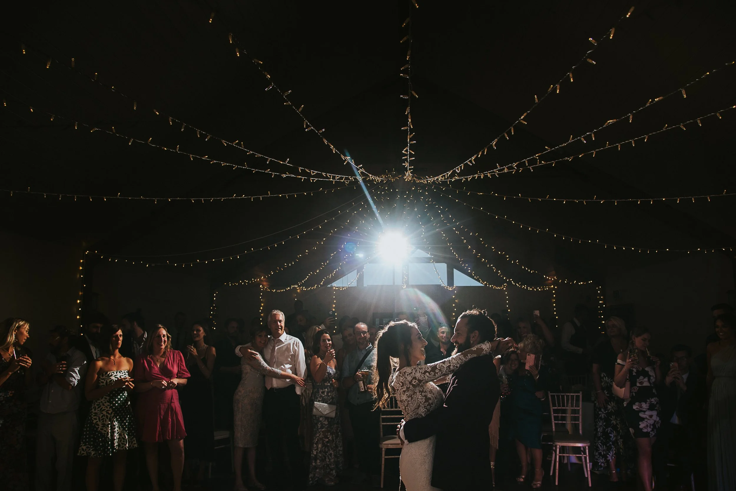 Bride and groom share first dance at wedding reception under fairy lights, surrounded by guests.