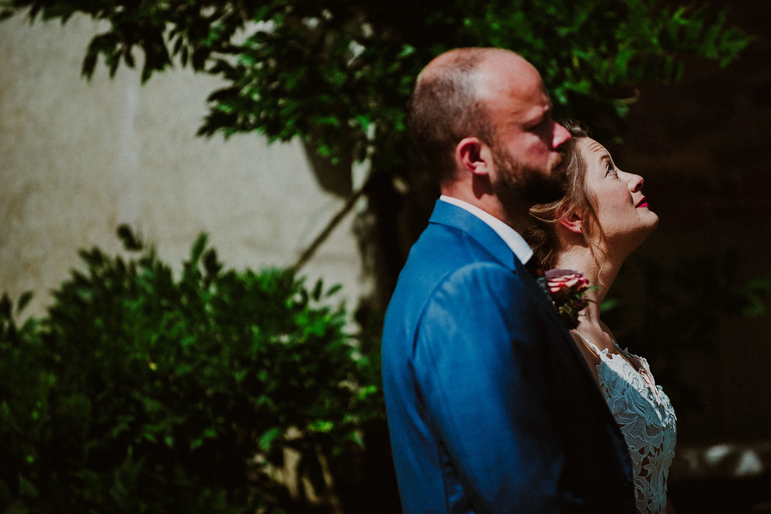 Couple in formal attire outdoors, man in blue suit, woman in white dress, looking up, with greenery background.