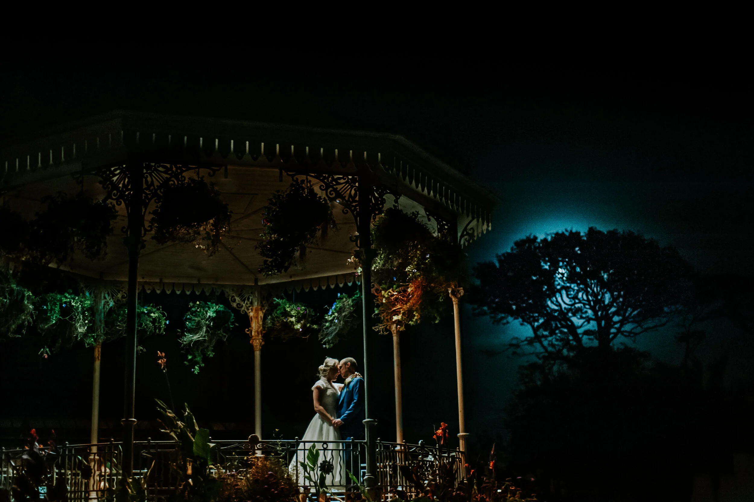Bride and groom embracing under illuminated gazebo at night with scenic backdrop