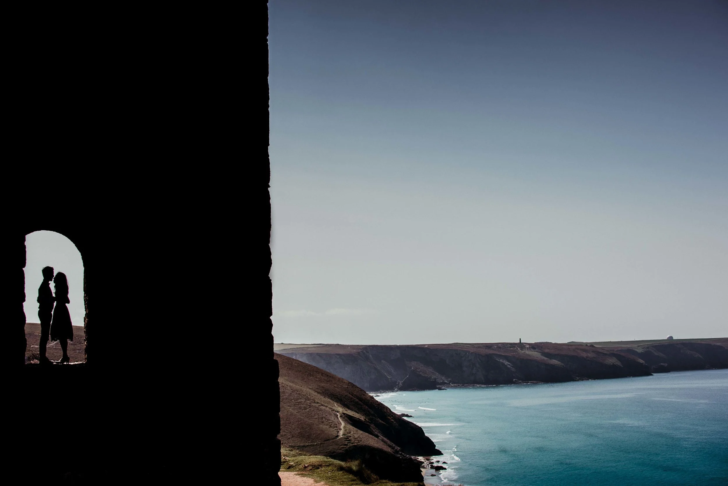 Silhouette of a wedding couple standing in the archway of an old Cornish mine, overlooking a coastal landscape with cliffs and the sea at Wheal Coates in Cornwall.