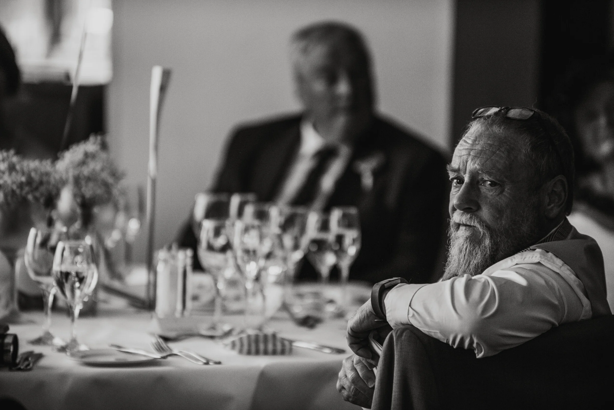 Black and white photo of a man with a beard sitting at a table with glasses and flowers, other people in the background, captured by award-winning Cornwall wedding photographer, Mark Shaw. 