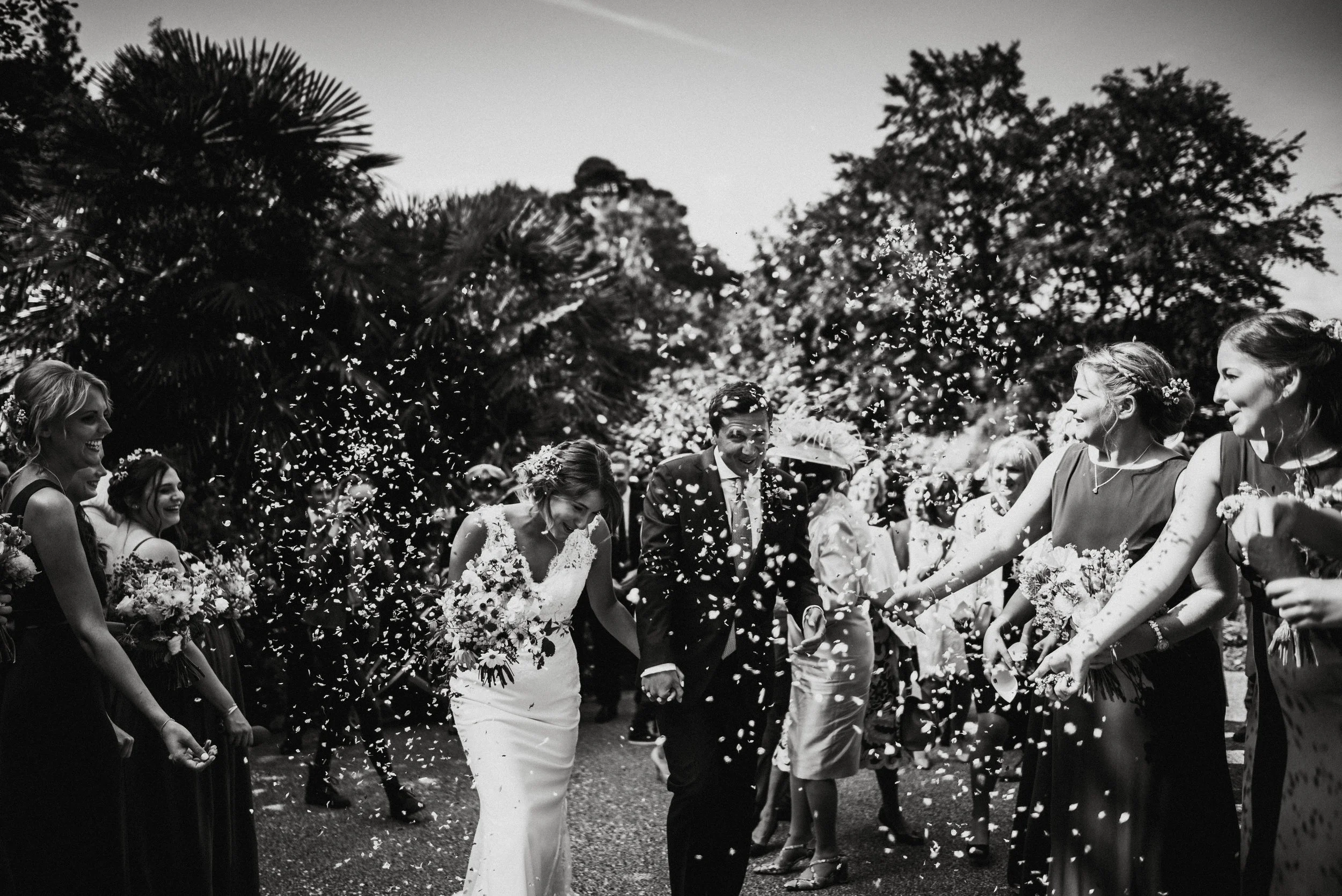 Black and white photo of a wedding celebration with a bride and groom walking through a shower of flower petals or confetti, surrounded by friends and family outside with trees in the background.