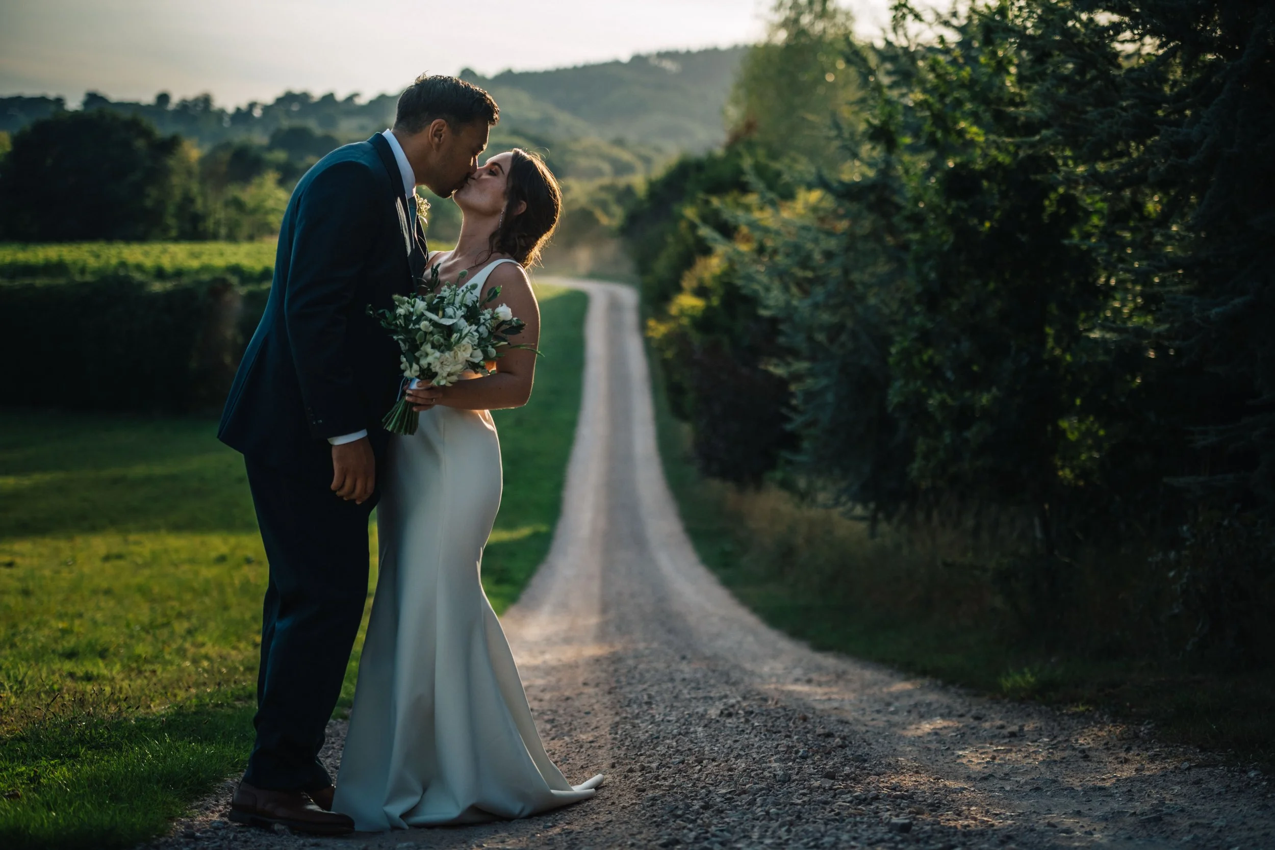 Couple kissing on a countryside path during sunset, woman holding bouquet, wedding attire, greenery background.