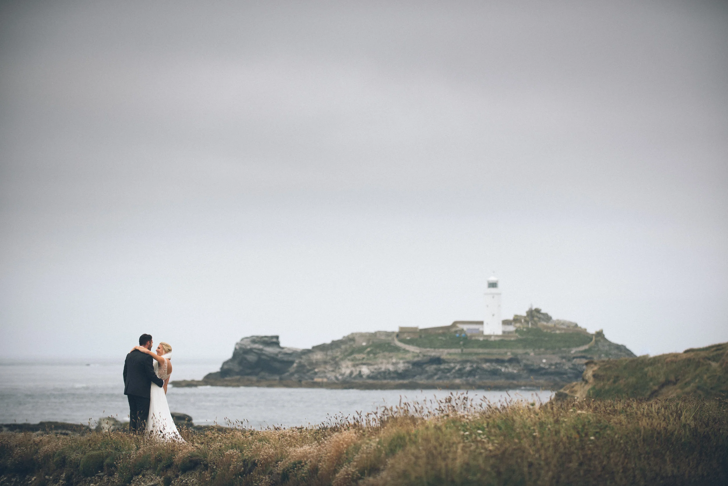 A newlywed couple embracing near a grassy shoreline, with a lighthouse on a rocky island in the background under a cloudy sky.