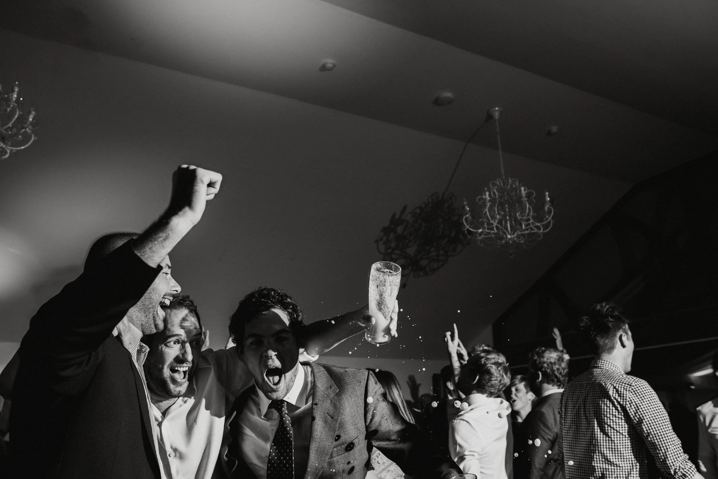 Black and white photo of people celebrating at a party, with one person holding a drink and others raising their arms, captured by award-winning Cornwall wedding photographer, Mark Shaw. 