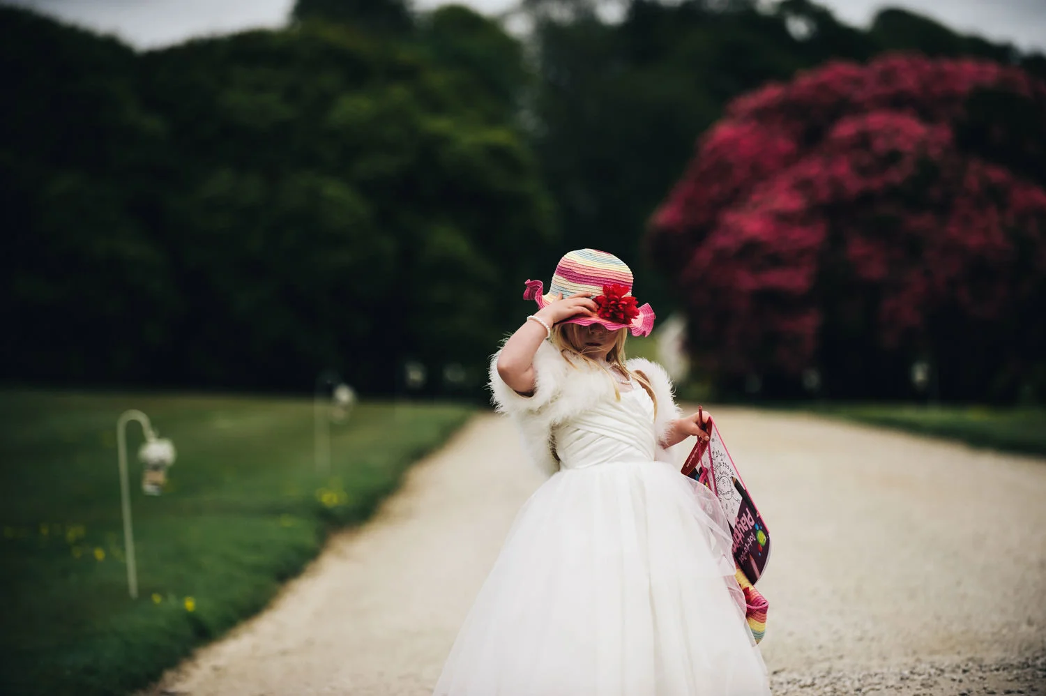 A young girl in a white dress and furry shawl wearing a wide-brimmed striped hat decorated with a pink flower, standing on a gravel pathway holding a purse and looking down with her hand adjusting the hat in a garden or park with green trees and a la